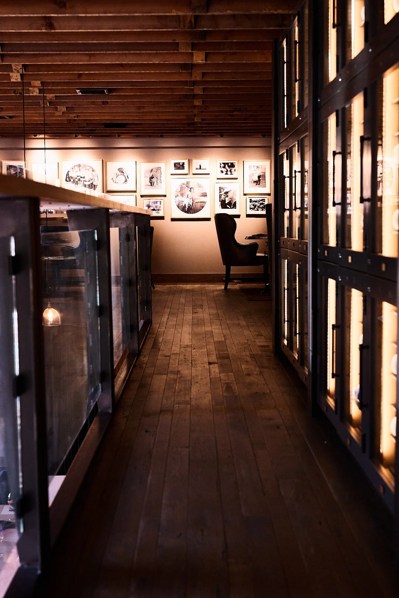 A cozy cafe or gallery with framed black-and-white photos on the wall, a wooden floor, and comfortable chairs, viewed through a glass and wood partition.