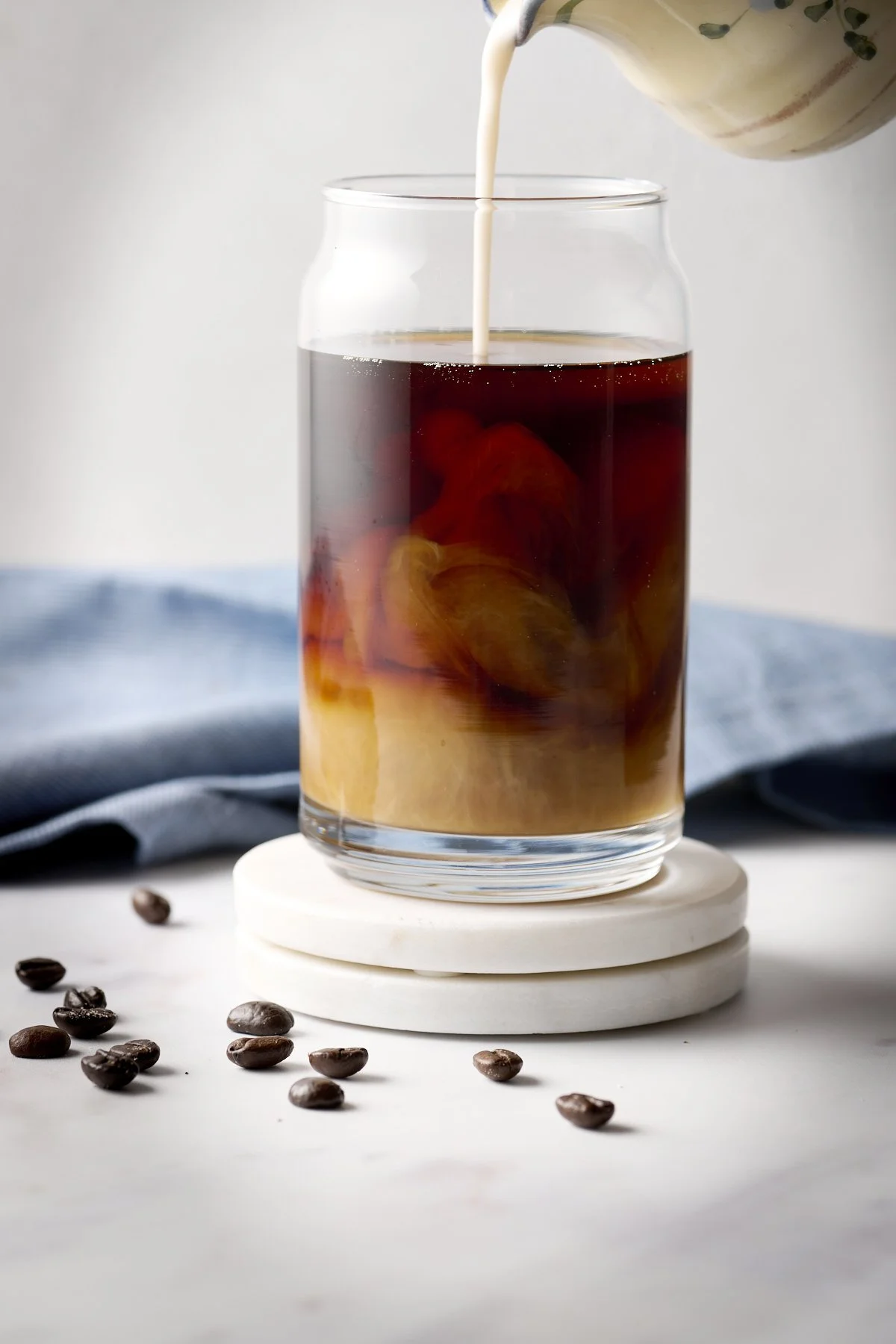A glass of iced coffee with cream being poured in, surrounded by scattered coffee beans, on a white surface with a blue cloth in the background.