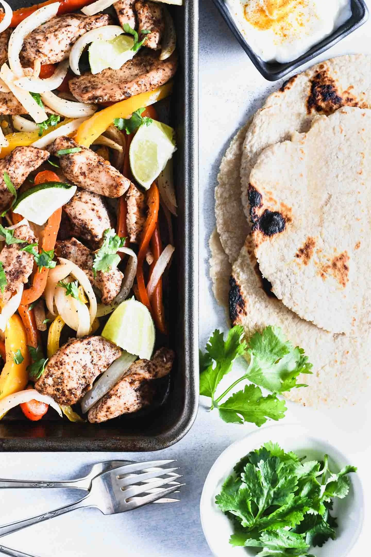 Grilled chicken fajitas with colorful peppers and onions, lime wedges on a metal tray, with saltine crackers, small bowl of sour cream, and a side bowl of fresh cilantro on a white surface.
