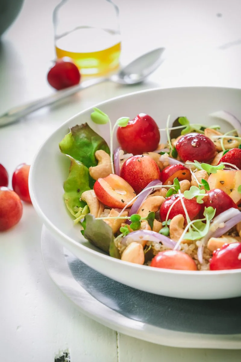 A bowl of salad with cherry tomatoes, mixed greens, red onion slices, cashews, and microgreens, with a spoon and a small glass container of salad dressing in the background.