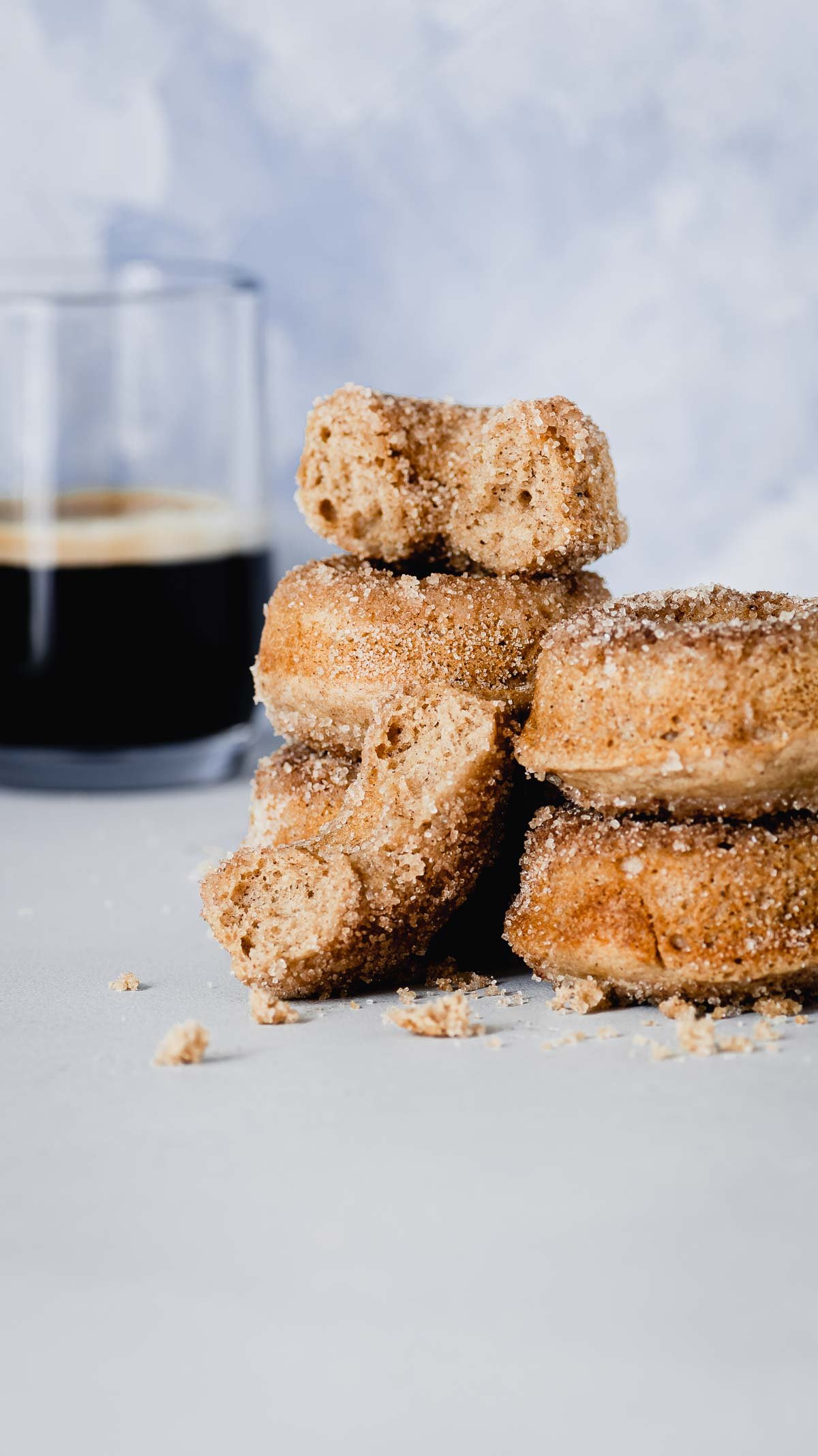A stack of donuts with a glass of dark coffee in the background on a white surface.