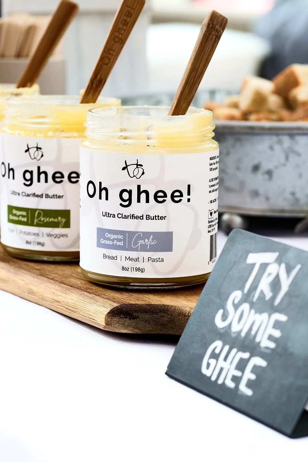Two jars of Oh Gee! Ultra Clarified Butter, one labeled garlic flavor, placed on a wooden board with a handwritten sign reading "Try some ghee" next to them.