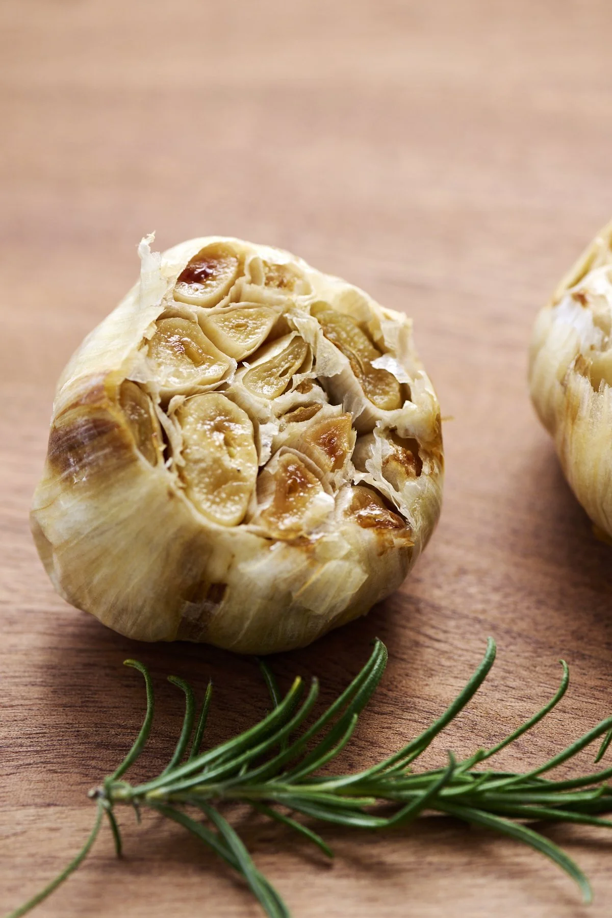 Close-up of roasted garlic bulb sliced open, showing individual cloves, on a wooden surface with a sprig of rosemary in front.