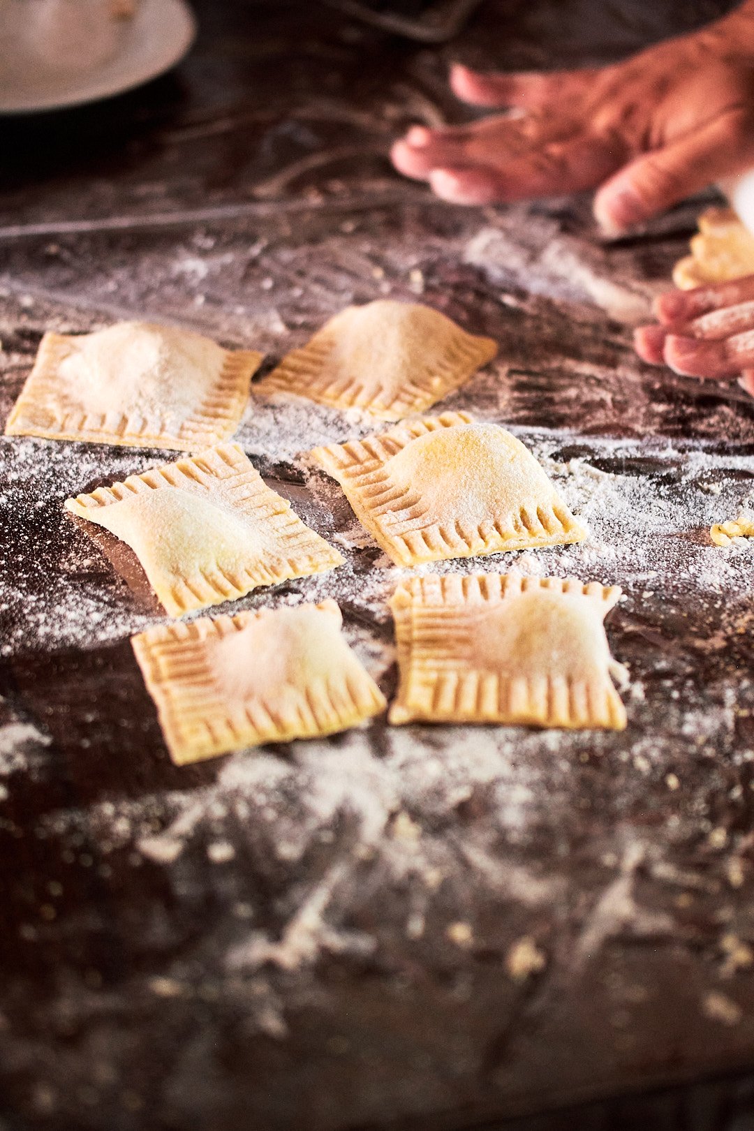 Uncooked ravioli pasta on a floured dark surface with a person's hands in the background.