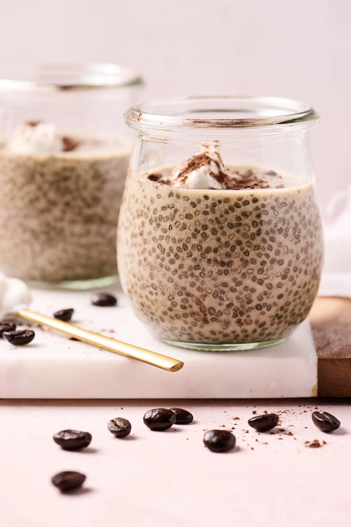 Close-up of two glass jars filled with chia pudding topped with whipped cream, with coffee beans scattered around on a pink surface.