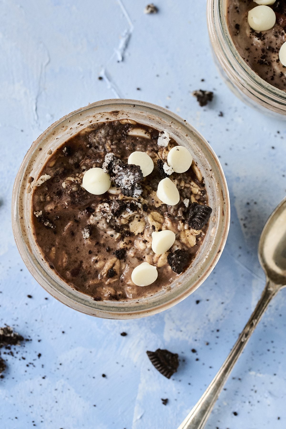 Close-up of a jar of chocolate overnight oats topped with white chocolate chips and crumbled cookie pieces, with scattered cookie pieces and a spoon on a light blue textured surface.