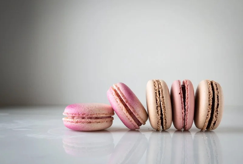 Five pink and beige macarons lined up on a white reflective surface against a plain light gray background.