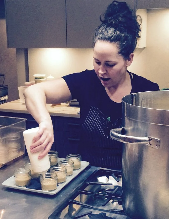 A woman in a black shirt is pouring a liquid into small glass jars on a tray in a kitchen.