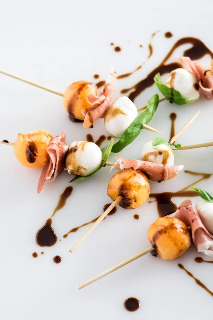 Skewers with mozzarella balls, cherry tomatoes, prosciutto, basil leaves, and balsamic glaze on a white plate.