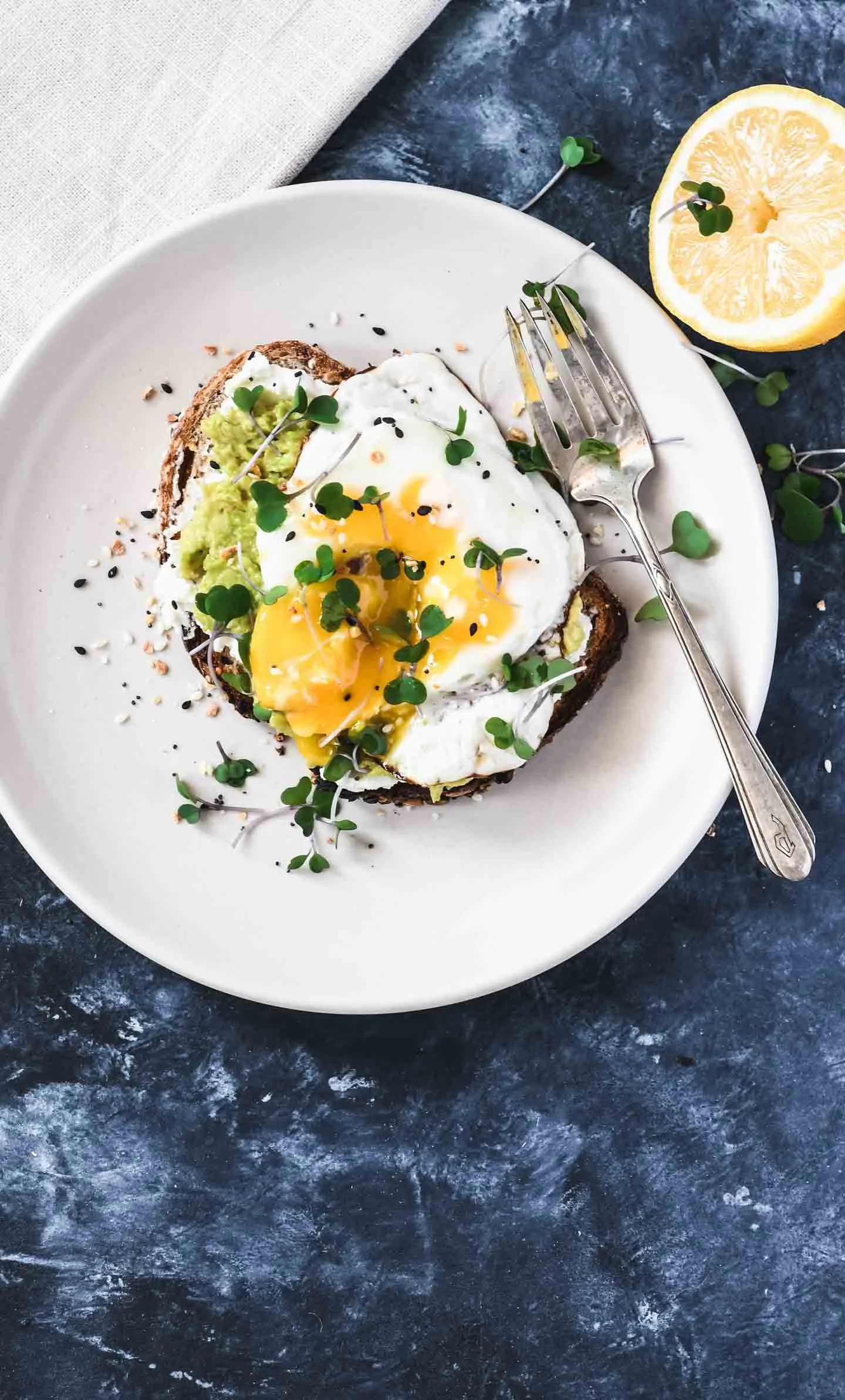 A white plate holds toast topped with mashed avocado, a fried egg with runny yolk, and microgreens. A silver fork rests on the plate, and a lemon half and microgreens are beside it. The setting includes a dark textured surface and a cream-colored nap