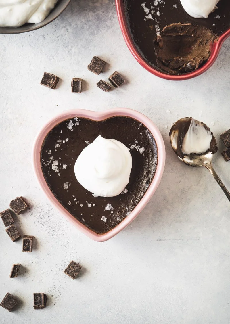Heart-shaped bowl of chocolate pudding topped with whipped cream and sea salt, with more pudding in a red bowl and chocolate cubes on a light surface.