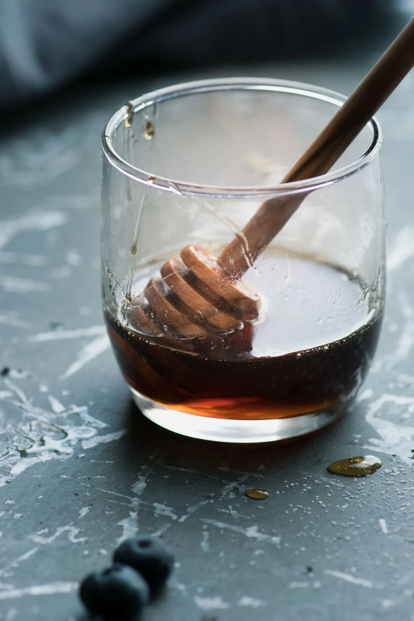 A glass bowl with honey and a honey dipper resting inside, placed on a dark textured surface with some honey drips and two blueberries in the foreground.