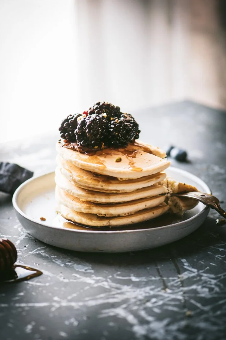 Stack of five pancakes topped with blackberries and syrup on a white plate.