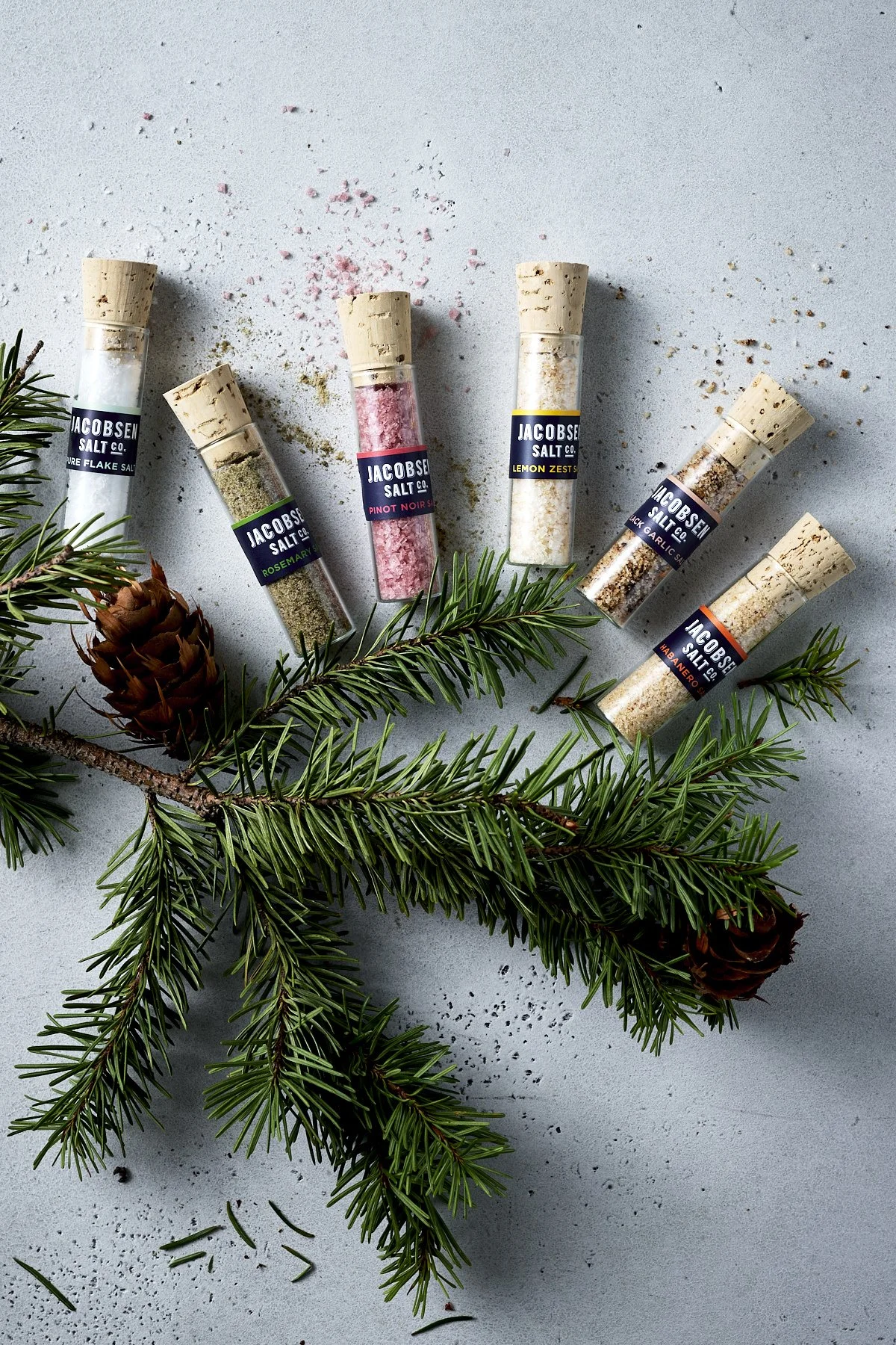 Six glass tubes of flavored salts arranged on a light gray surface with small scattered salt crystals and a pine branch with pinecones.