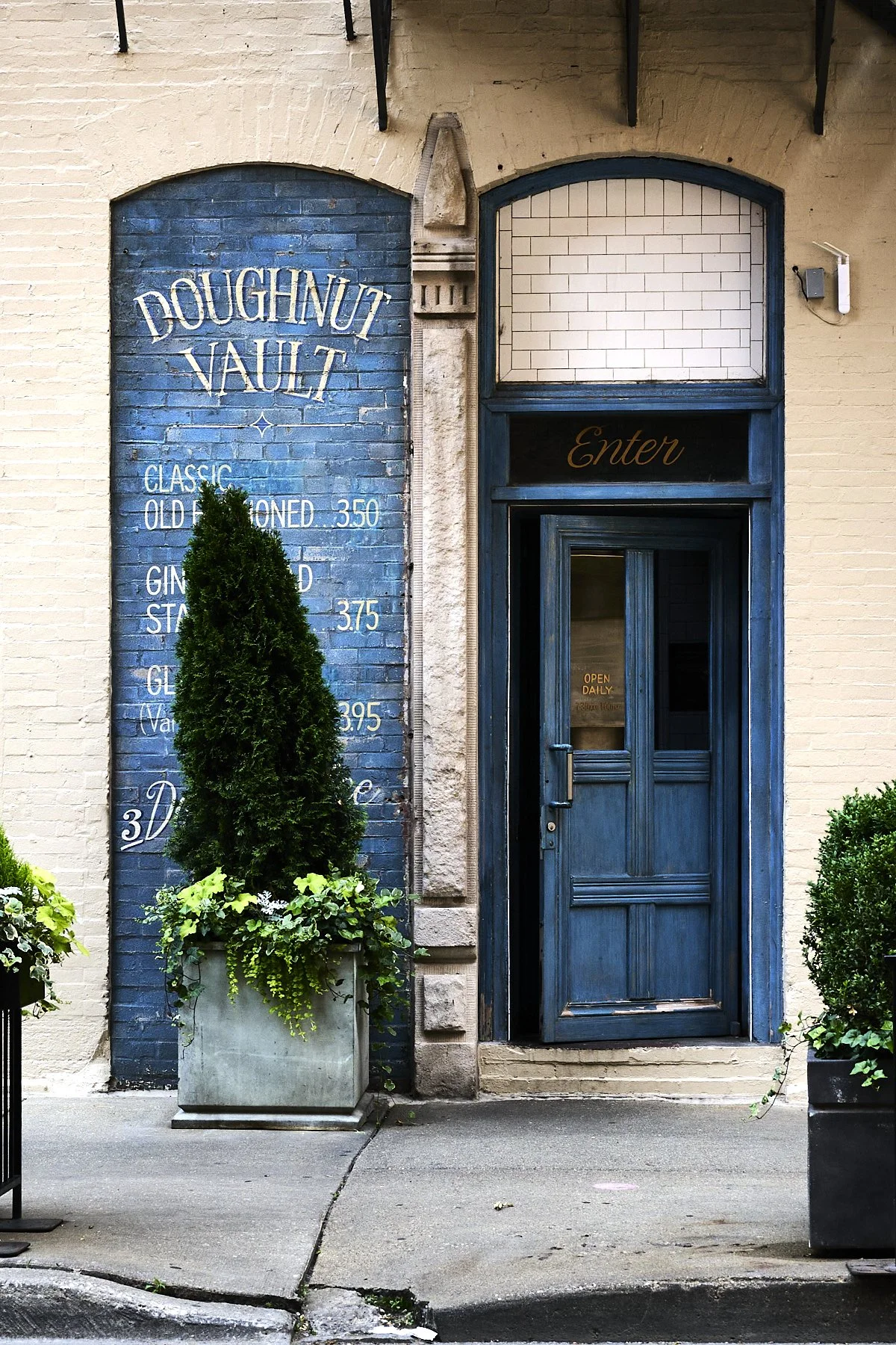 Exterior of a bakery with a blue door, a sign reading "Doughnut Vault," and potted plants outside.
