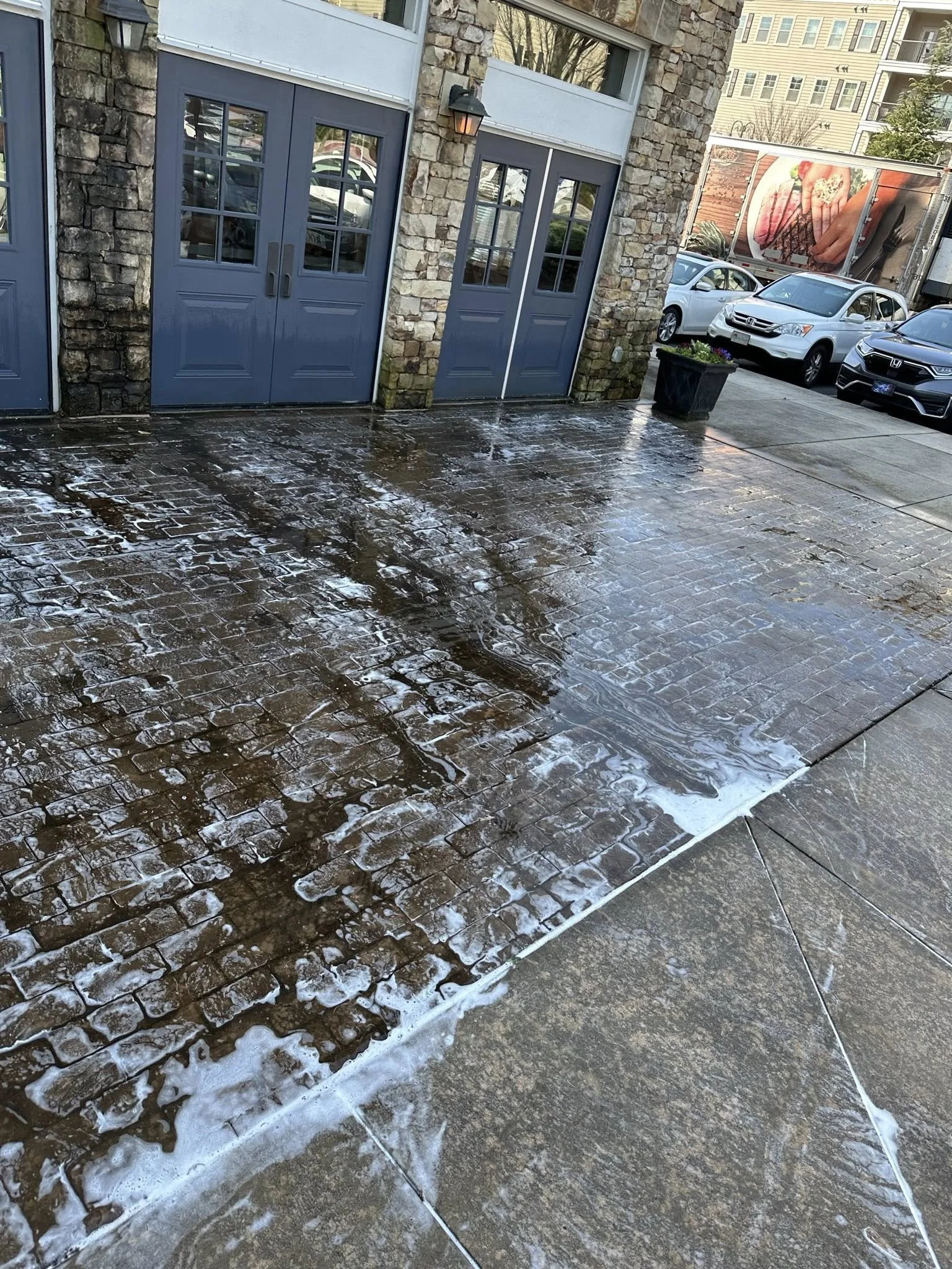 Wet brick and concrete driveway with soap suds or cleaning foam, outside a building with blue doors, stone and brick exterior, next to parked cars, in an urban setting.
