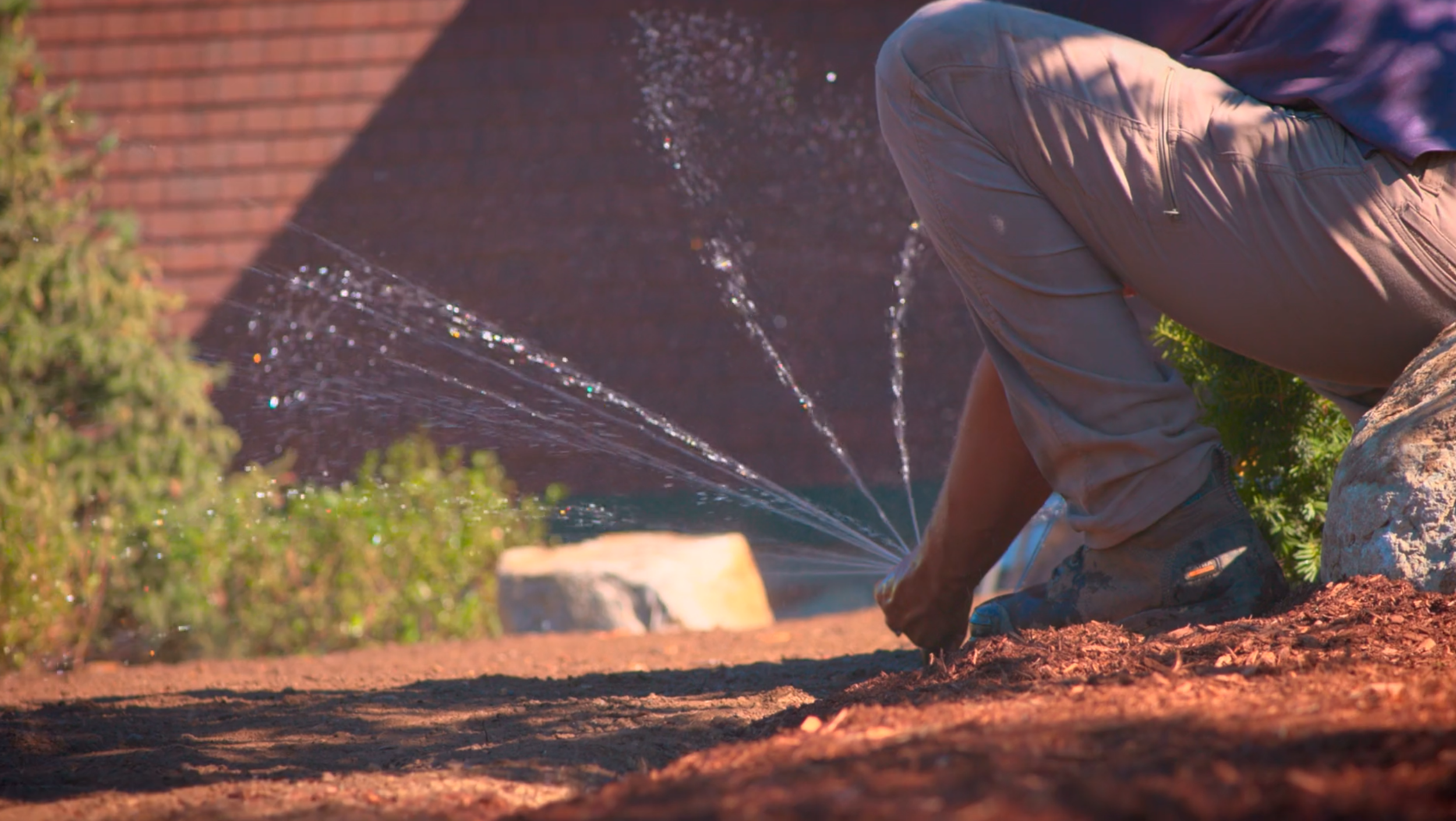 A person kneeling on the ground adjusting irrigation heads during the daytime.