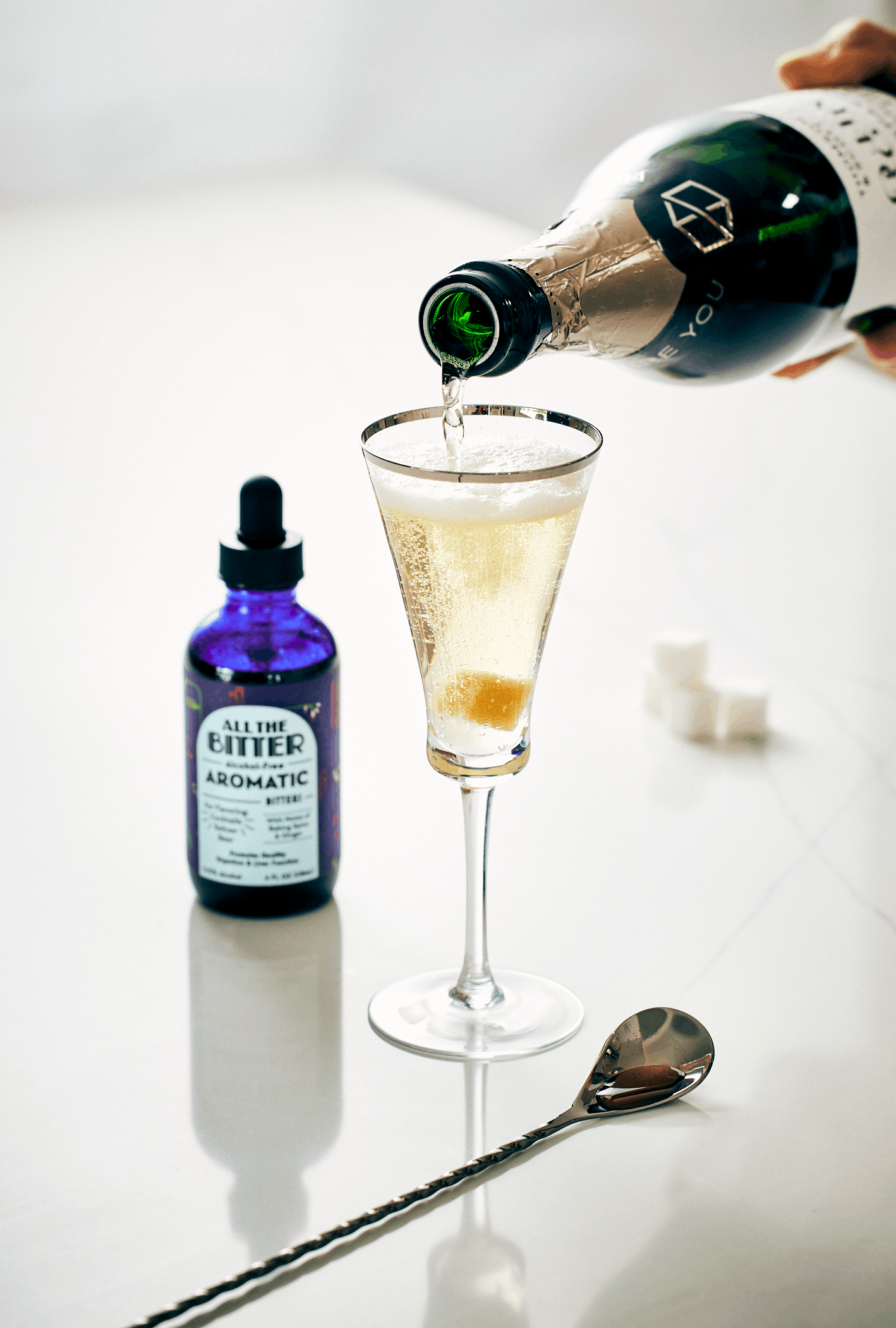 A hand pouring champagne into a martini glass with an orange slice, with a bottle labeled "All the Bitter Aromatic Bitters" and a silver cocktail spoon on a white table.