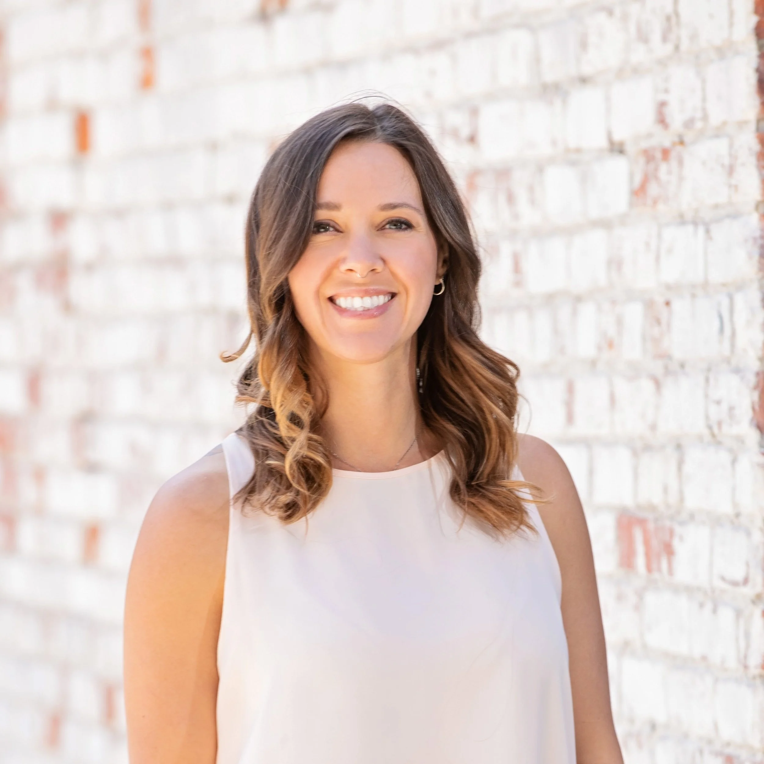 Portrait of a smiling woman with brown wavy hair standing against a white brick wall.