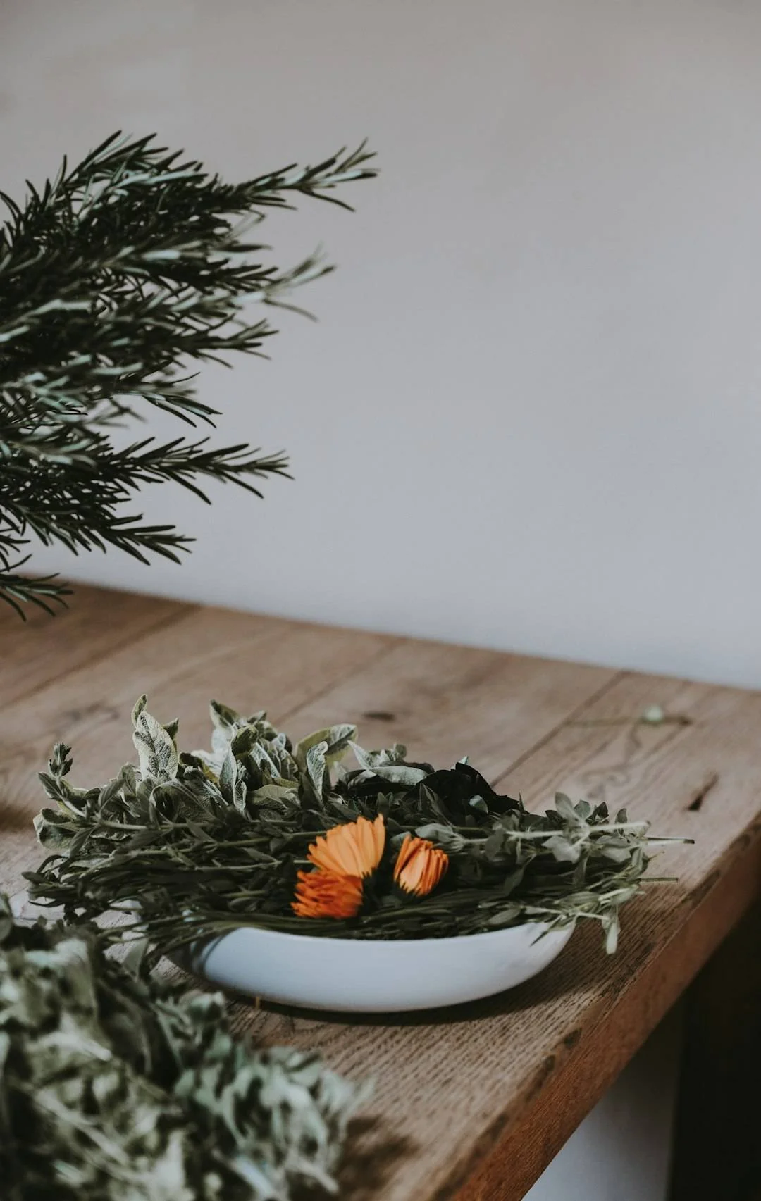 A white oval ceramic bowl filled with green foliage, orange flowers, and grayish leaves, placed on a wooden surface against a plain light-colored background.