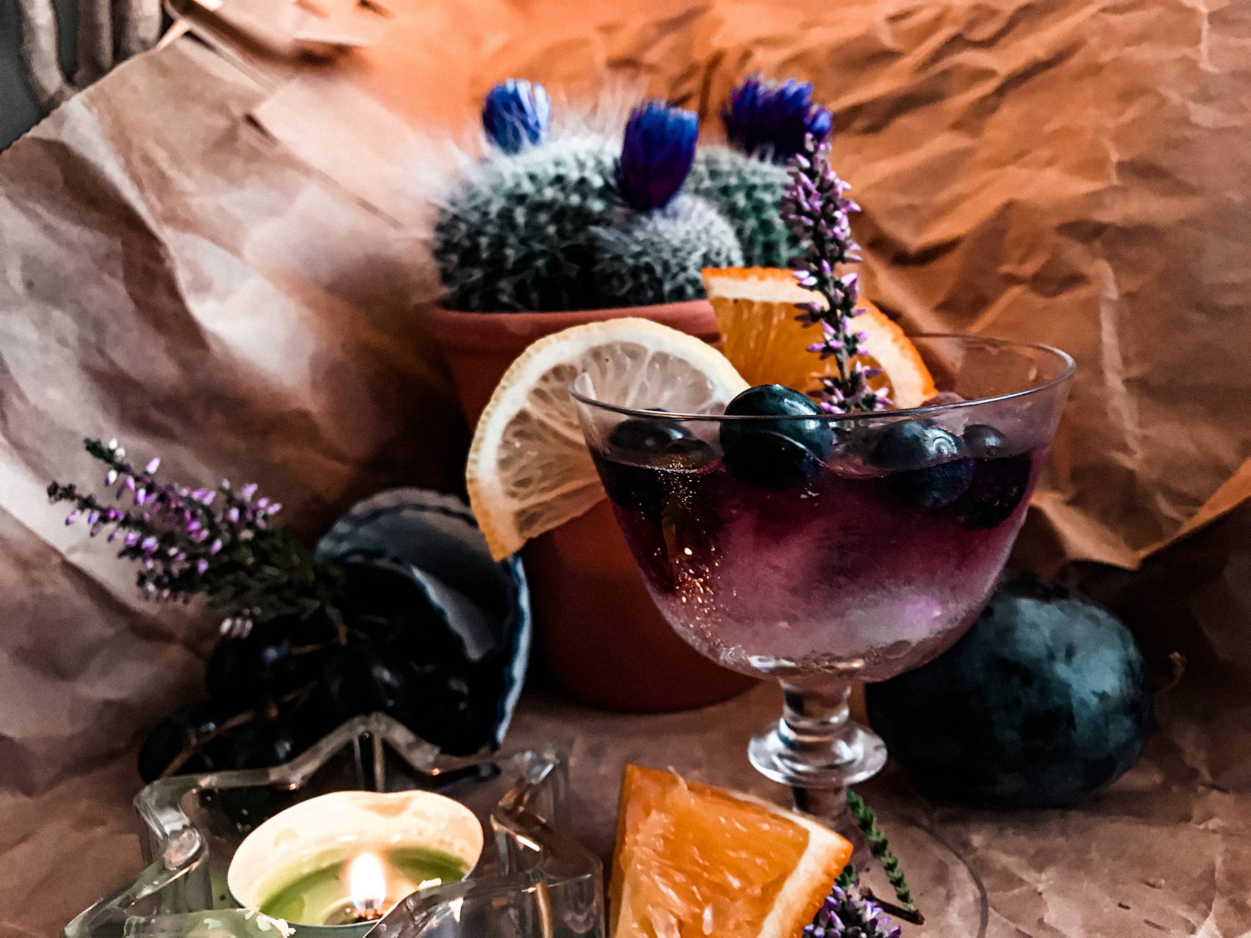 A glass of dark purple wine with blueberries, garnished with a sprig of purple flowers and slice of lemon, surrounded by oranges, a cactus with purple flowers, grapes, and a candle, all on crumpled brown paper.