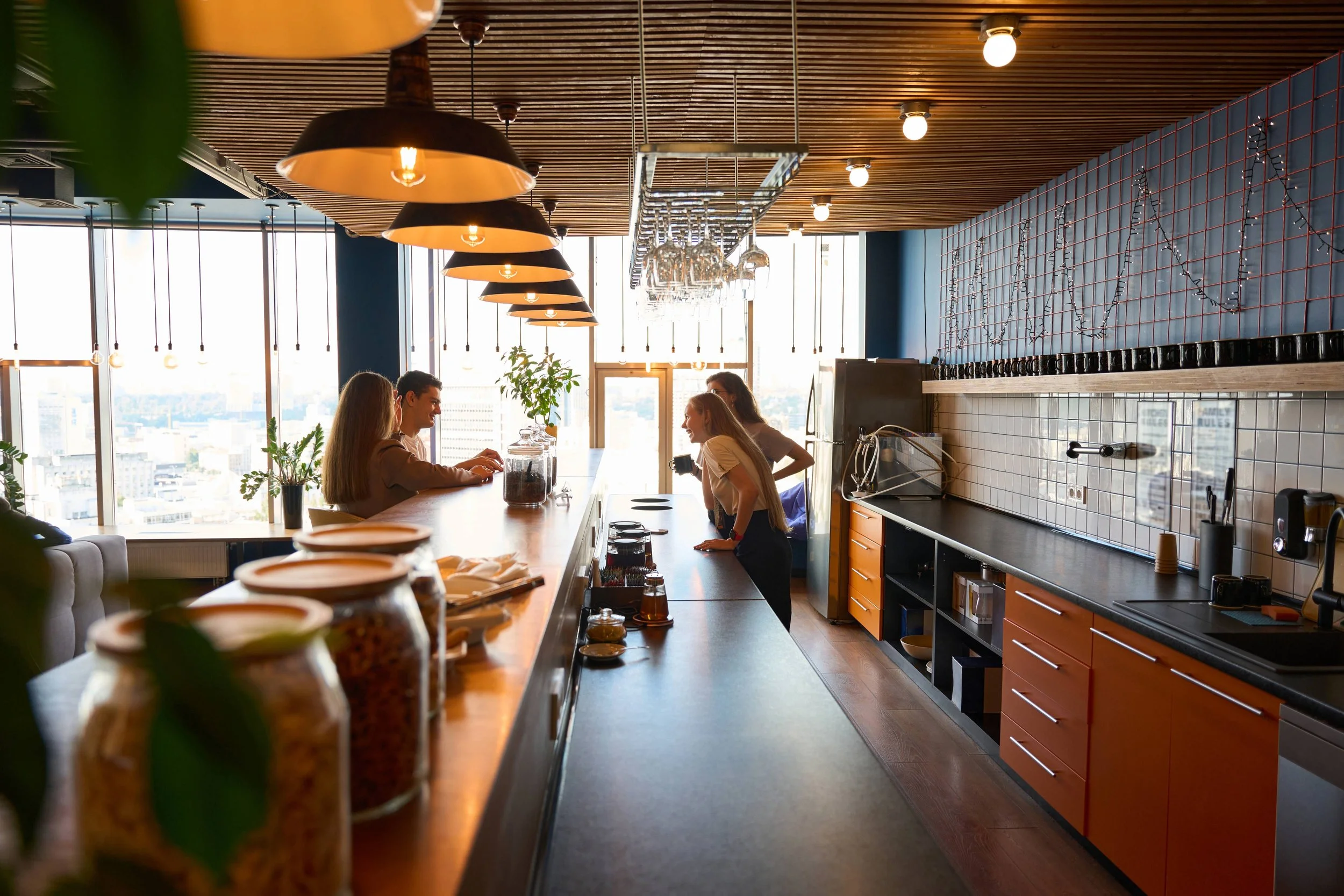 Group of people sitting and talking at a modern cafe or bar during daytime with large windows, plants, and stylish lighting.