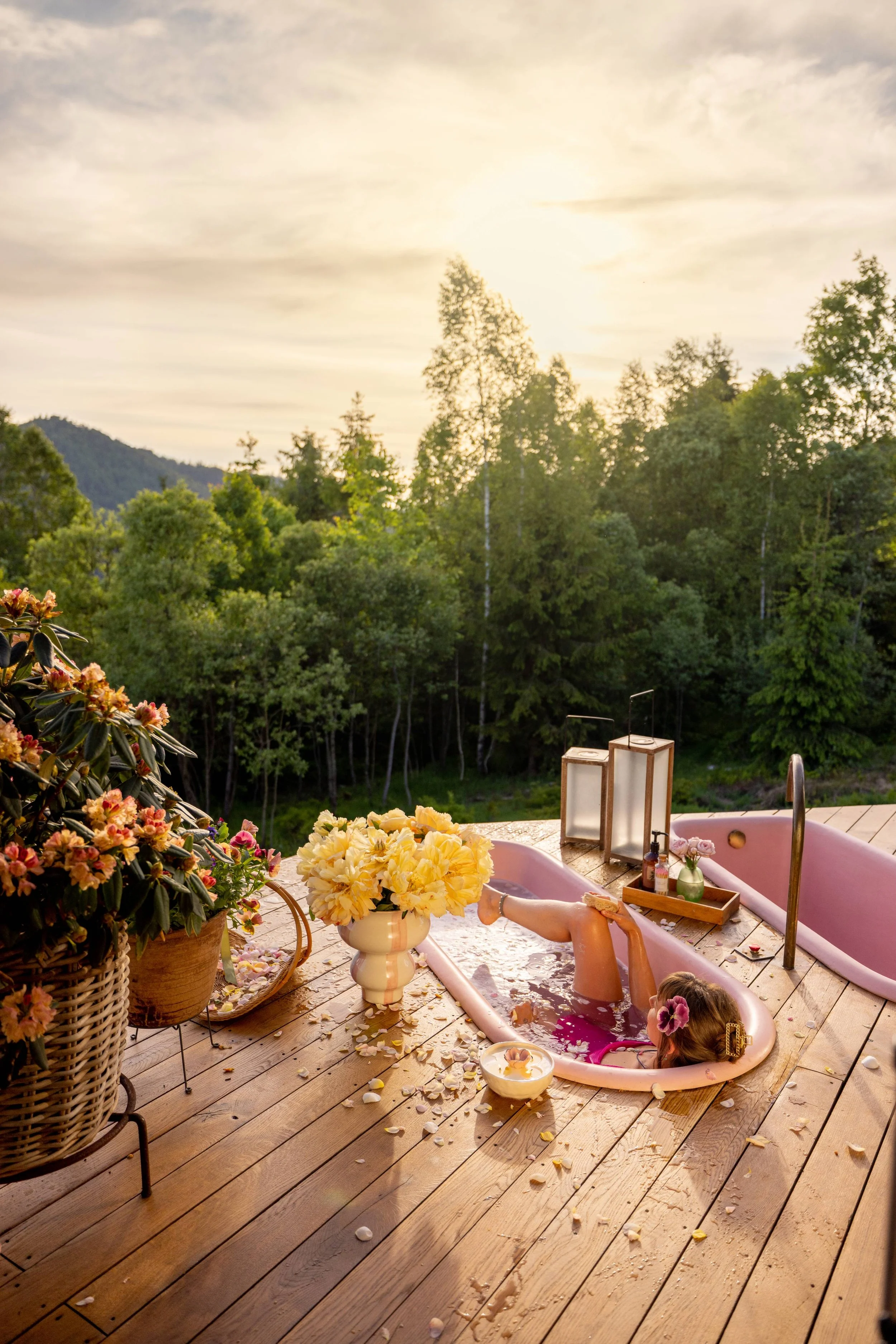 A girl relaxing in a pink bathtub on a wooden deck outdoors during sunset, surrounded by flowers and overlooking a green forest.