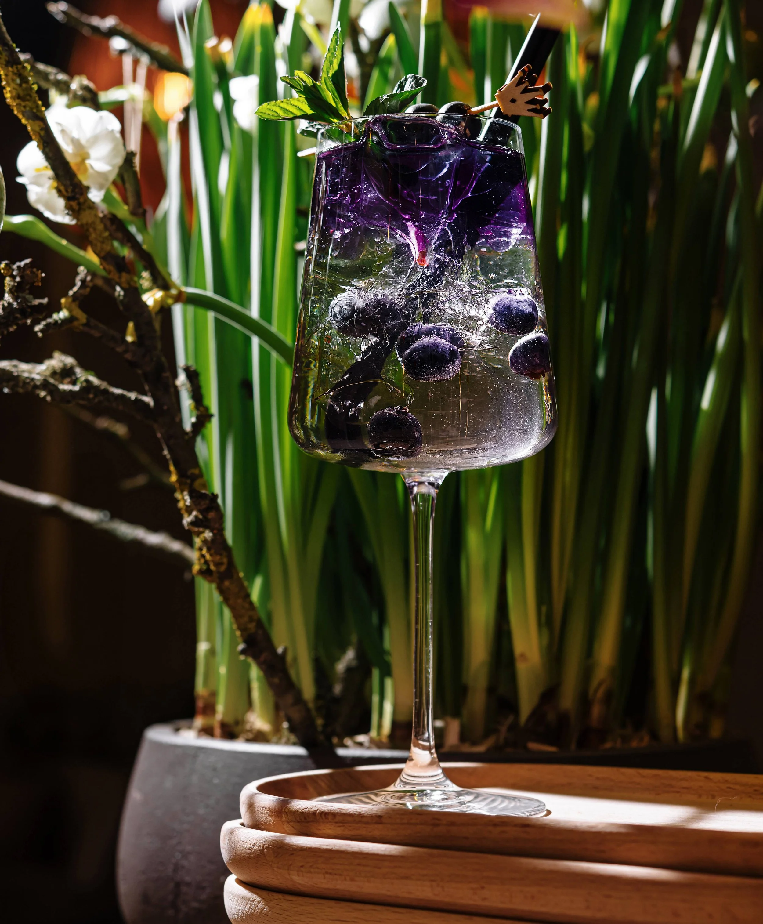 A tall wine glass filled with a purple-blue beverage, blueberries, ice, and garnished with mint and a decorative umbrella, placed on a wooden tray with green plants in the background.