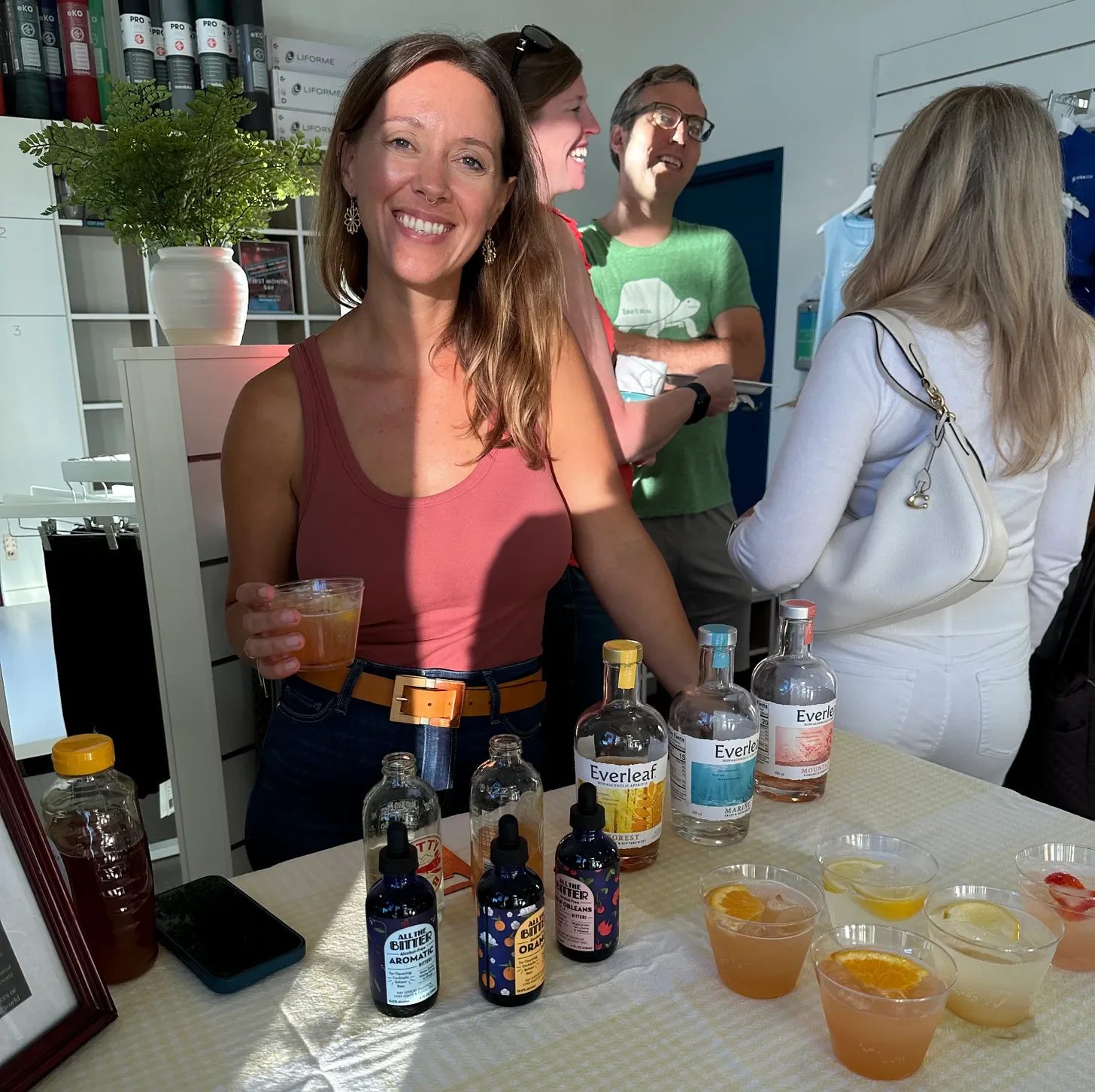A woman smiling and holding a drink at a booth with bottles and samples of flavored drinks on a table. People are in the background, and the setting appears to be an indoor event or good.