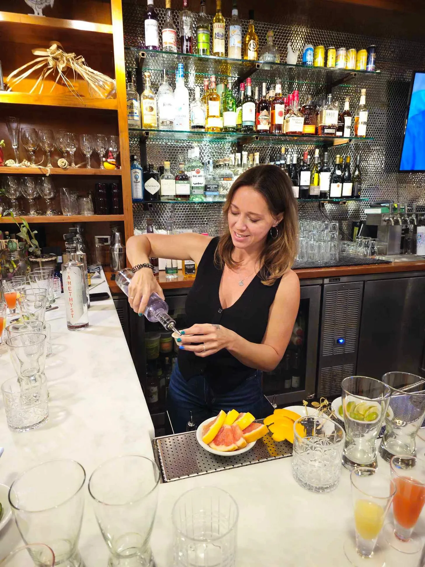 A woman preparing drinks behind a bar with various bottles of alcohol on shelves in the background, and a plate of sliced fruit on the bar counter.
