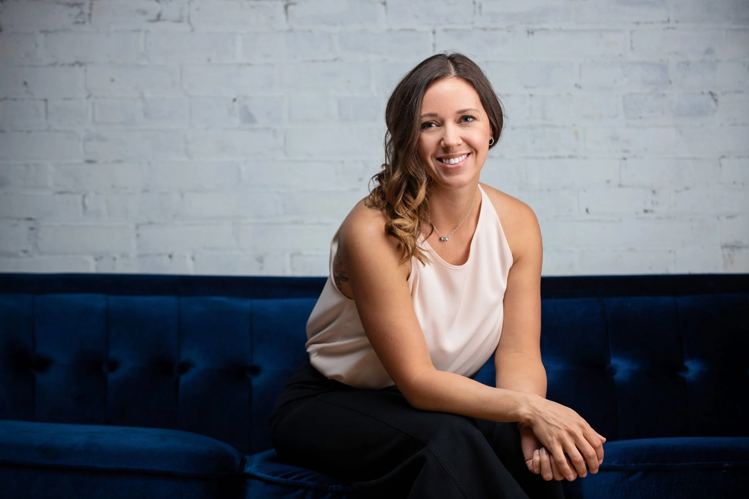 A smiling woman with wavy brown hair, wearing a beige sleeveless top, sitting on a dark blue velvet sofa with her hands clasped. She is against a white brick wall.
