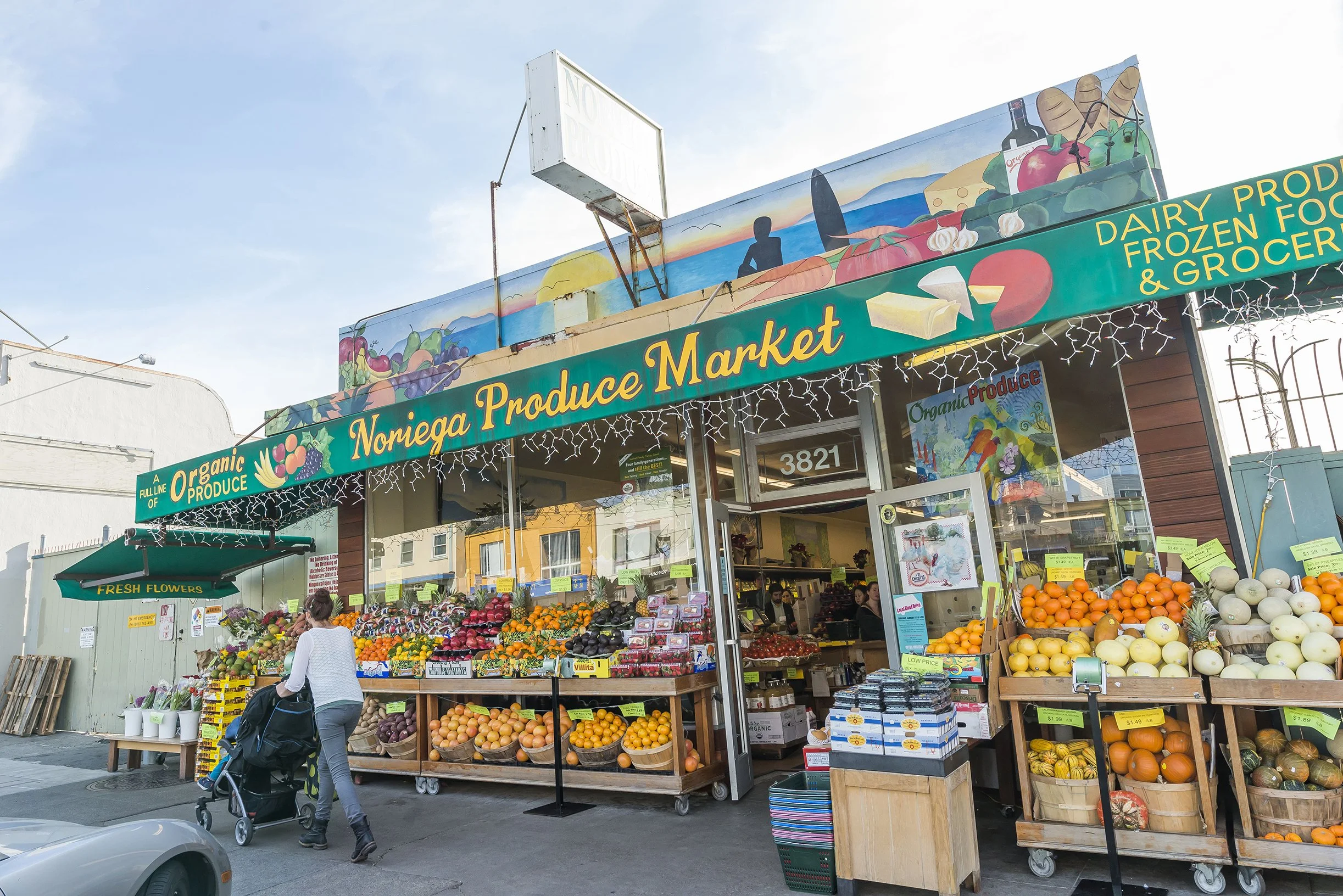 Front view of a produce market with fresh fruits and vegetables displayed outside, a woman with a stroller walking in front, and a store sign that reads "Noniega Produce Market."
