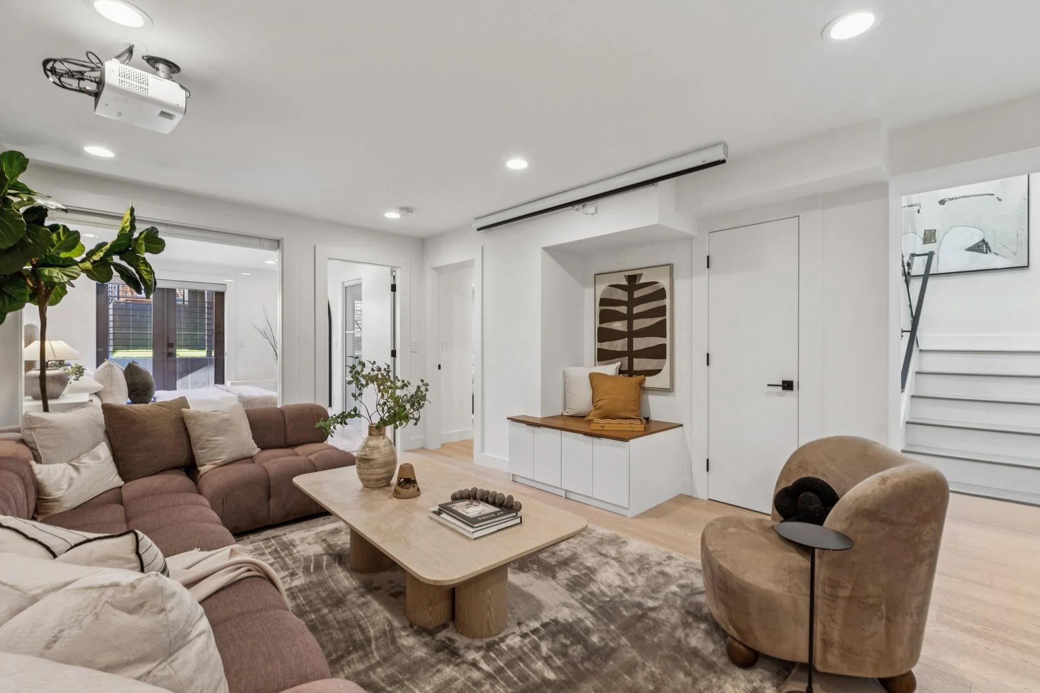 Living room with beige and brown furniture, a large sectional sofa, a wooden coffee table, a beige armchair, a rug, and indoor plants. There are windows and a staircase leading up.