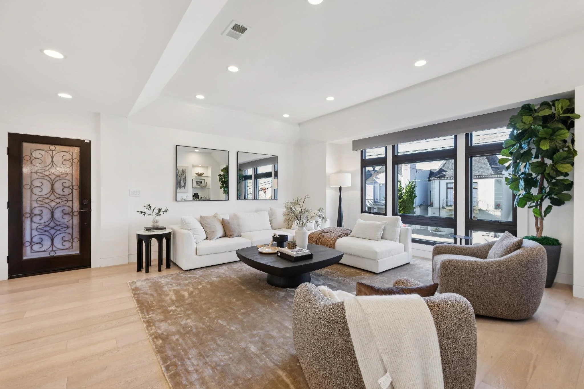 Modern living room with white sofas, a brown area rug, and large windows with black frames. There are two armchairs, a black coffee table, a tall floor lamp, and indoor plants. The walls are decorated with two mirrors and shelves with decorative items.