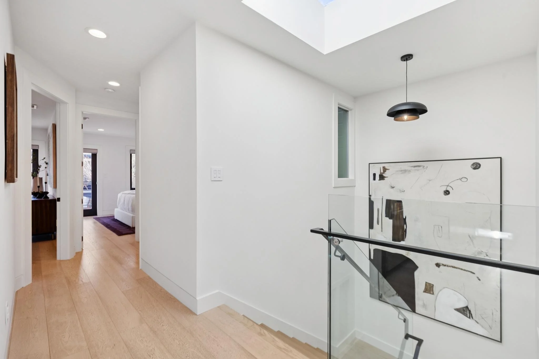 A hallway with light wood flooring leading to a bedroom, with white walls and ceiling. There is a black hanging light fixture and abstract artwork on the wall at the staircase landing.