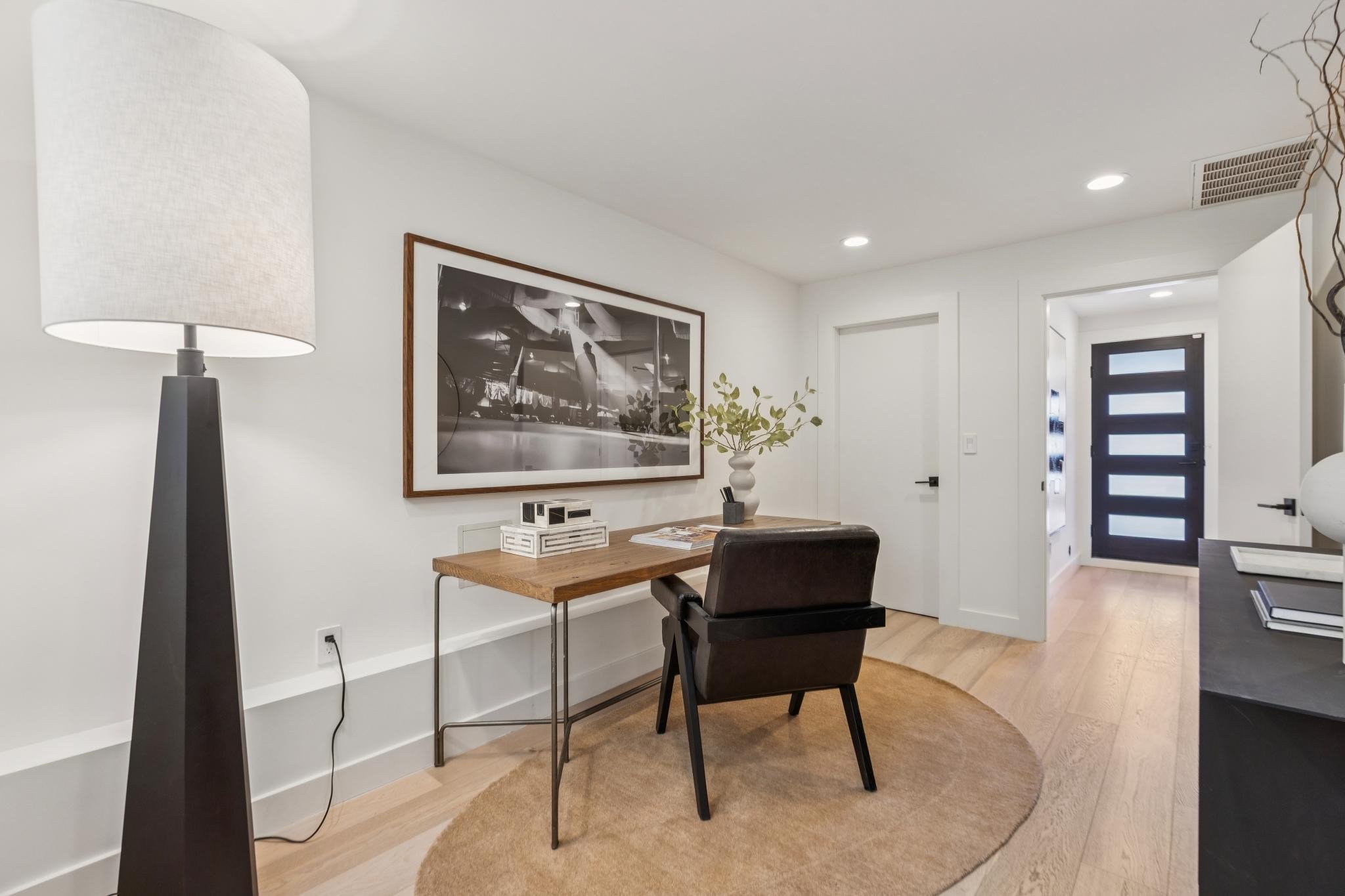 Modern entryway with a black front door with horizontal glass panels, white walls, wooden floor, a black sideboard with decorative items, a desk with a chair, a large black-and-white wall art, a potted plant, and a beige rug.