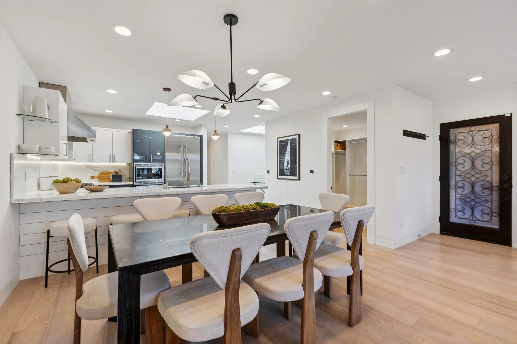 Modern open-concept kitchen and dining area with white walls, hardwood floors, a black dining table with beige upholstered chairs, a white kitchen island, stainless steel appliances, and black and white artwork on the wall.
