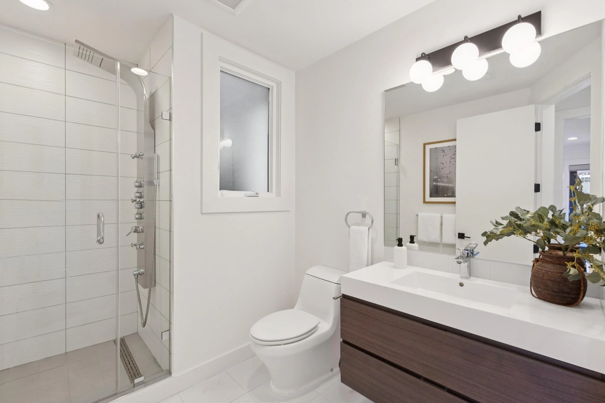 Modern white bathroom with a glass shower enclosure, a toilet, a wooden vanity with a white sink, a large mirror, a plant in a brown pot, and a window.
