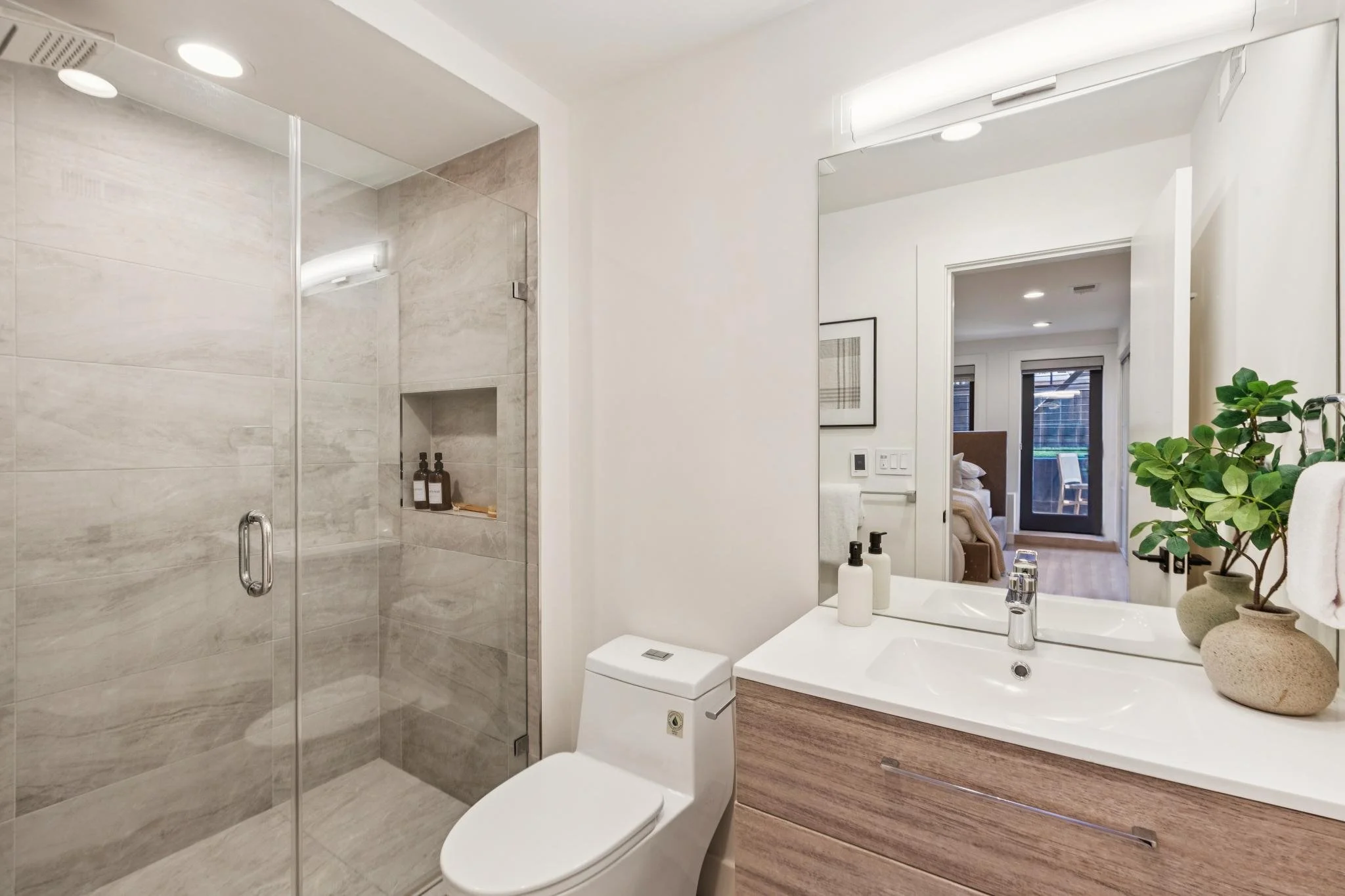 Modern bathroom with glass shower enclosure, white toilet, white countertop with vessel sink, large mirror, potted plant, and bedroom visible through open door.