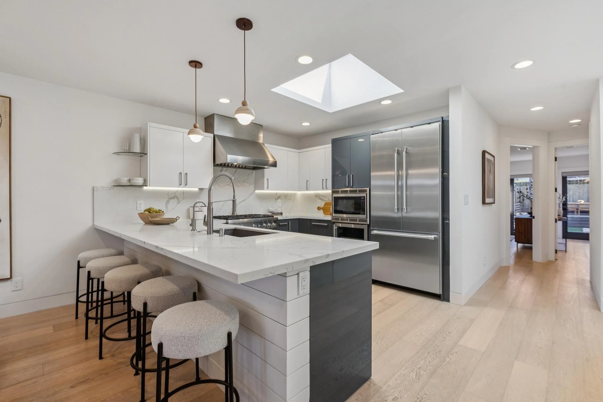 Modern kitchen with white and dark gray cabinets, stainless steel refrigerator, oven, microwave, and range hood, a kitchen island with a marble top, and four bar stools, with hardwood floors and a skylight.