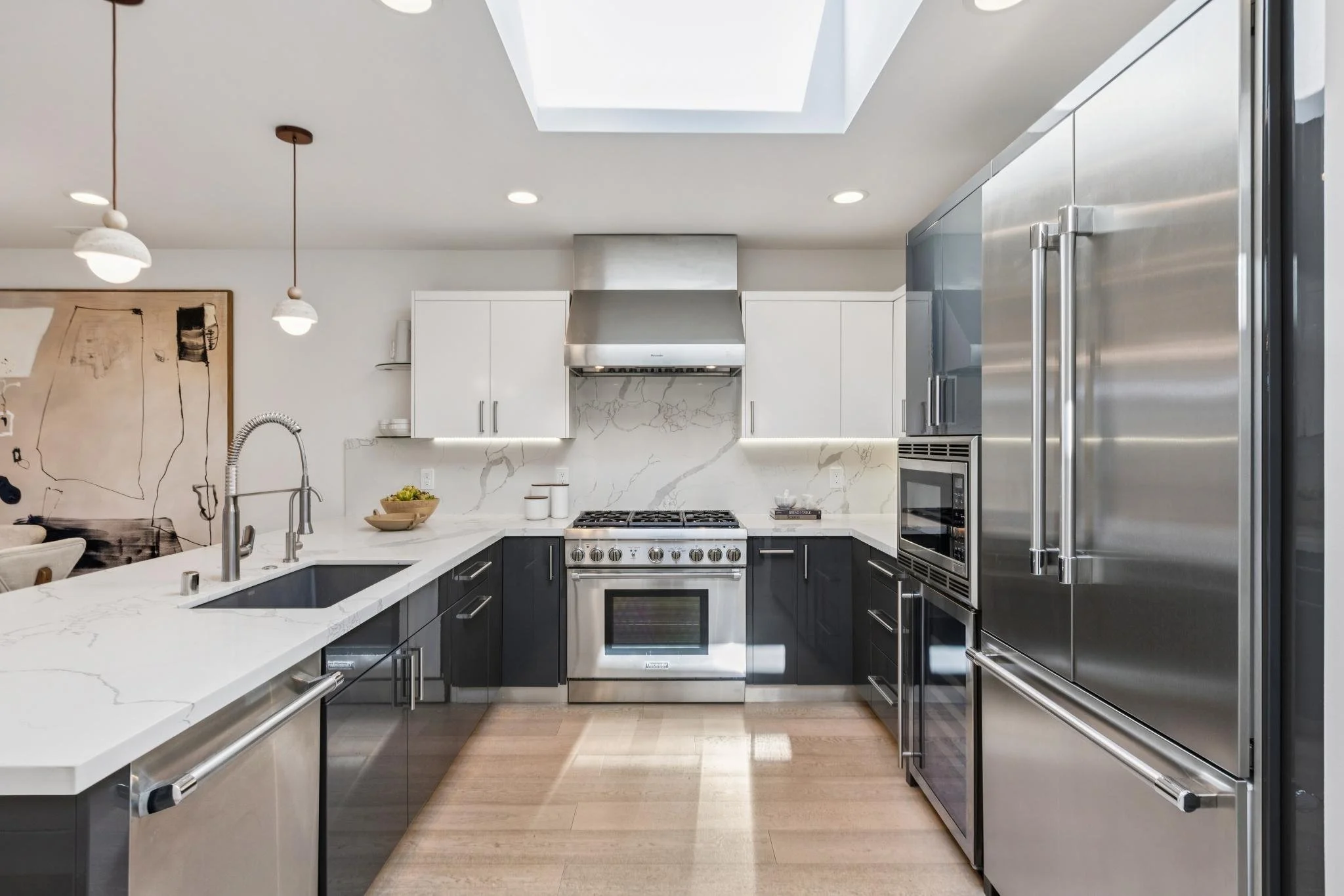 Modern kitchen with stainless steel appliances, white countertops, and black lower cabinets. Includes a large refrigerator, oven, microwave, and a skylight overhead.