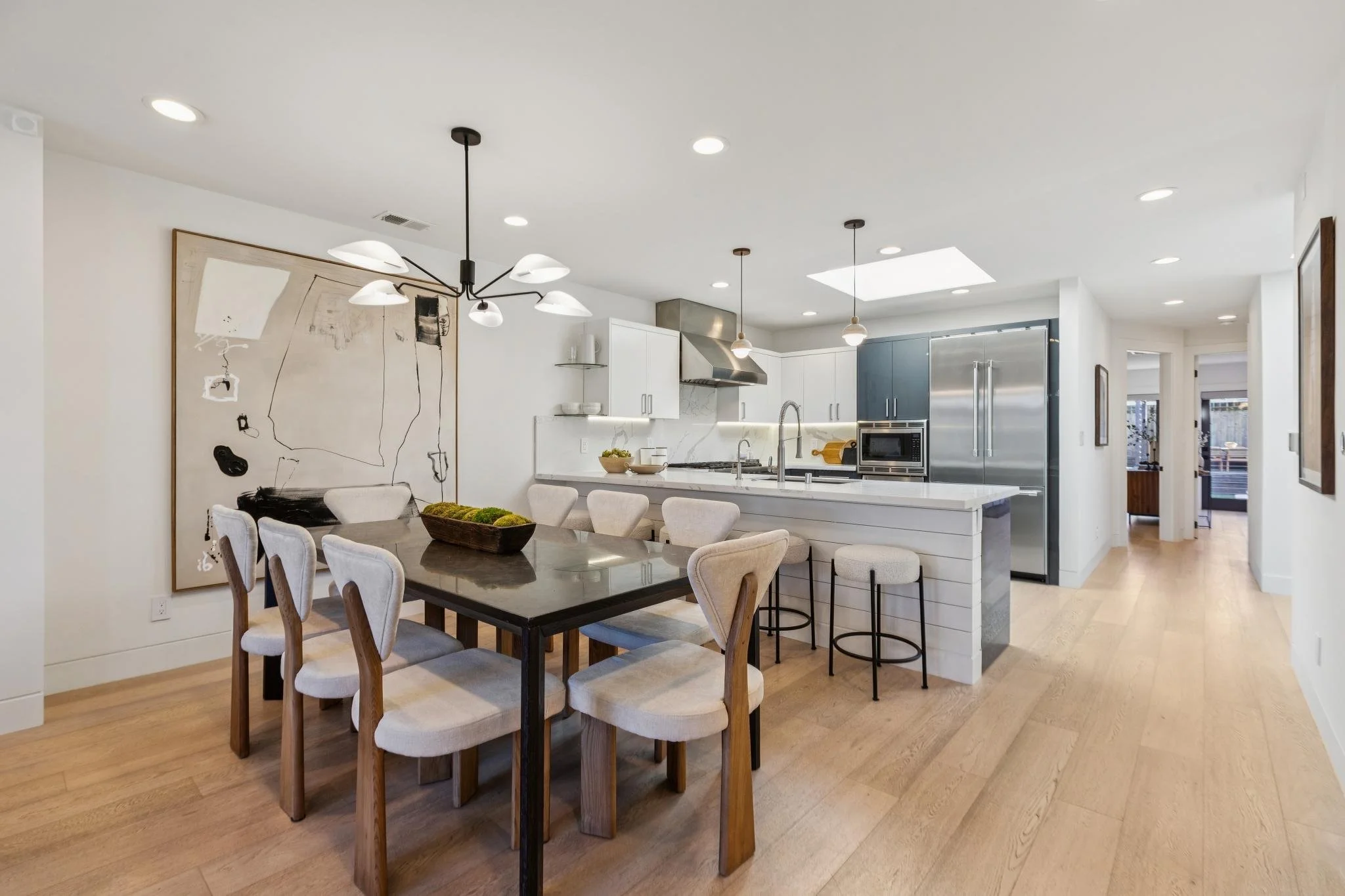Modern open-concept kitchen and dining area with wooden flooring, white walls, a black dining table with beige chairs, artwork on the wall, and kitchen appliances including a large refrigerator, oven, and microwave.
