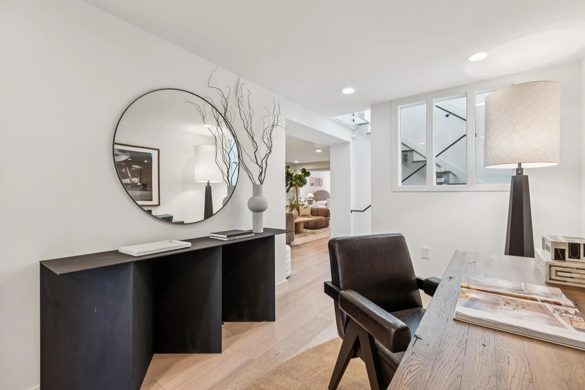 Living room with a black console table, a large round mirror, white walls, a table lamp, a black leather chair, a wooden dining table with magazines, and a staircase visible through a window.