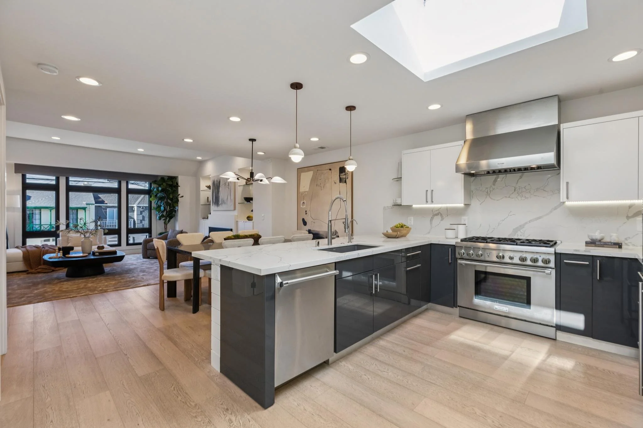 Modern open-concept kitchen with white and black cabinetry, marble countertops, stainless steel appliances, a large island with sink, and a living area with large windows and outdoor balcony doors.