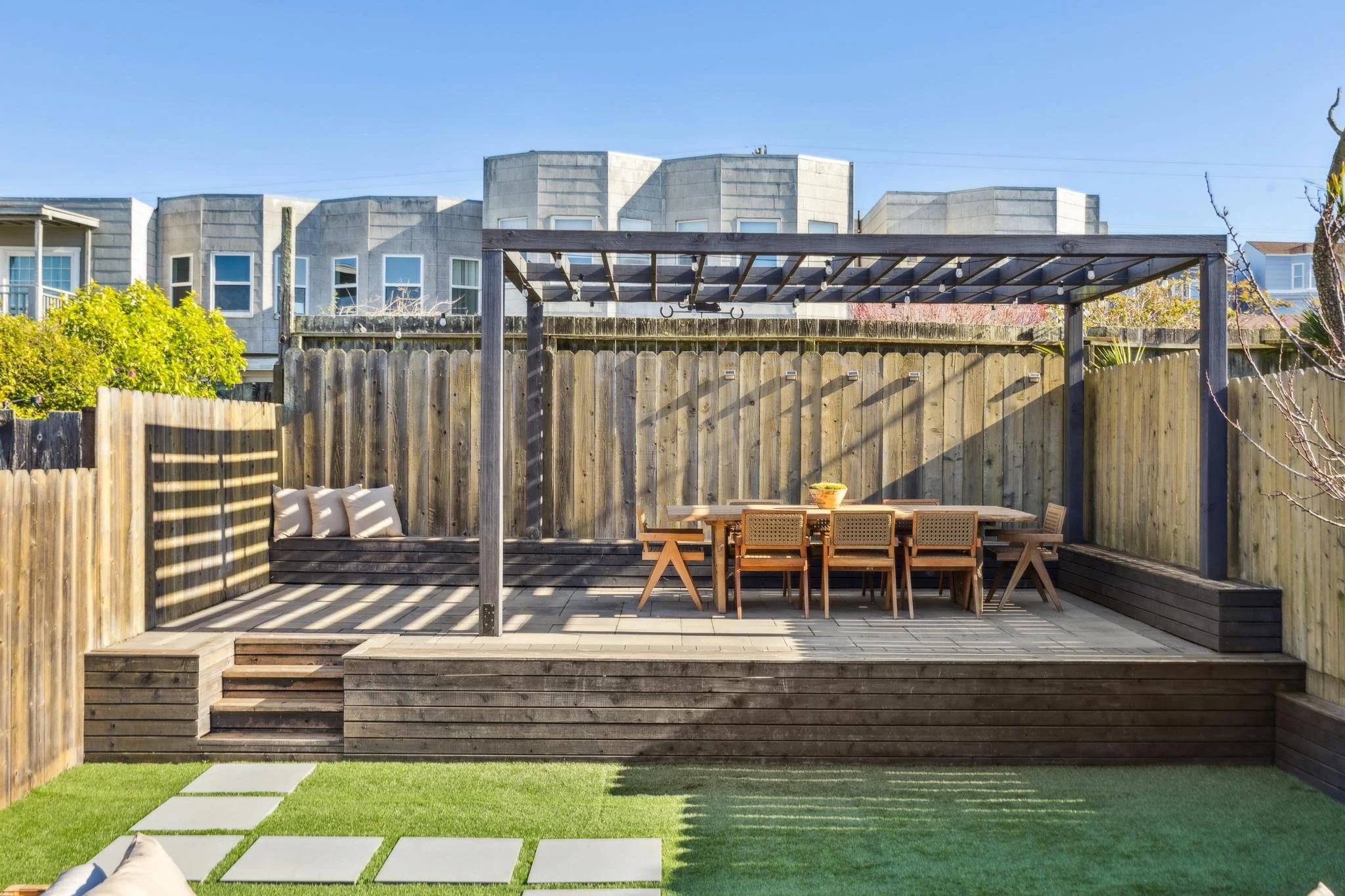 Backyard patio area with wooden deck, outdoor dining table with chairs under a pergola, and a grassy area with stone pavers.