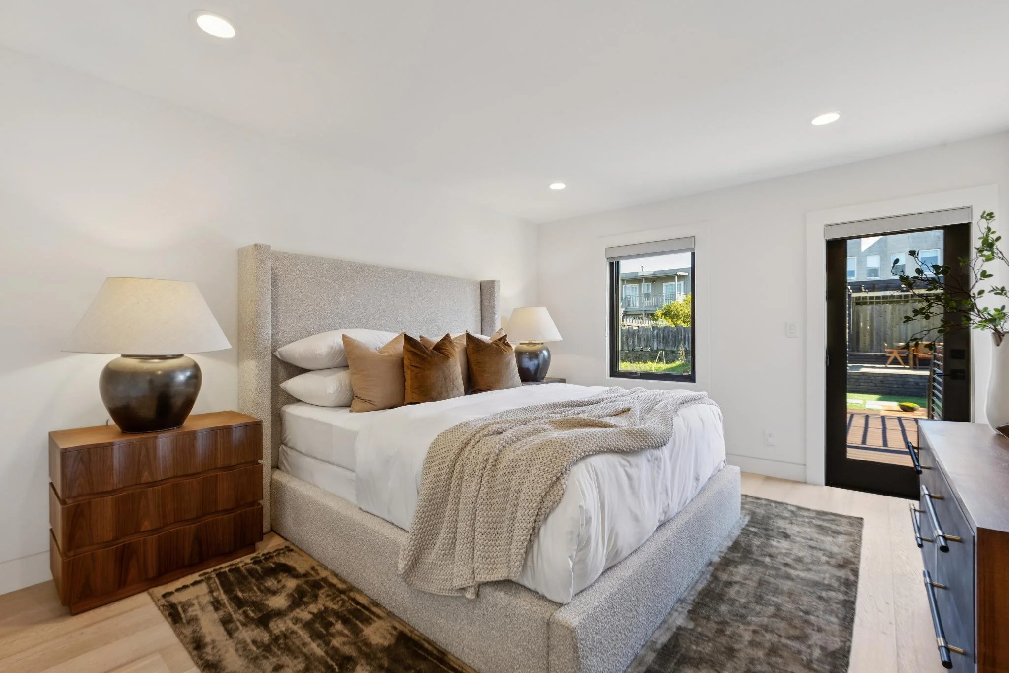 A bright bedroom with a large bed, beige headboard, white bedding, brown pillows, beige blanket, wooden nightstands with lamps, a window showing outdoor greenery, oak flooring, and a sliding door leading to a patio with outdoor seating.