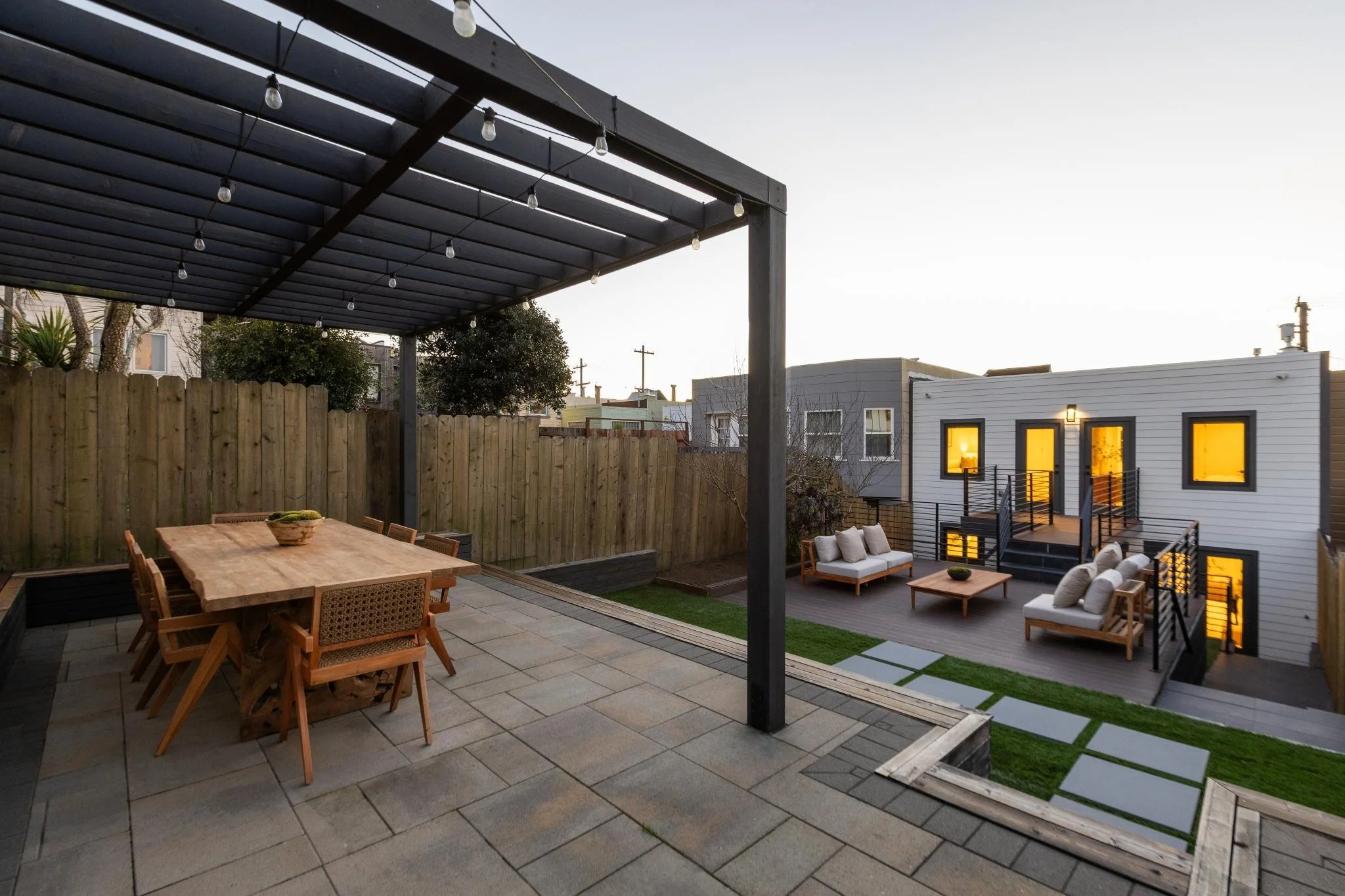 An outdoor patio with a wooden table and chairs under a black pergola with string lights, surrounded by a tall wooden fence and neighboring houses in the background.