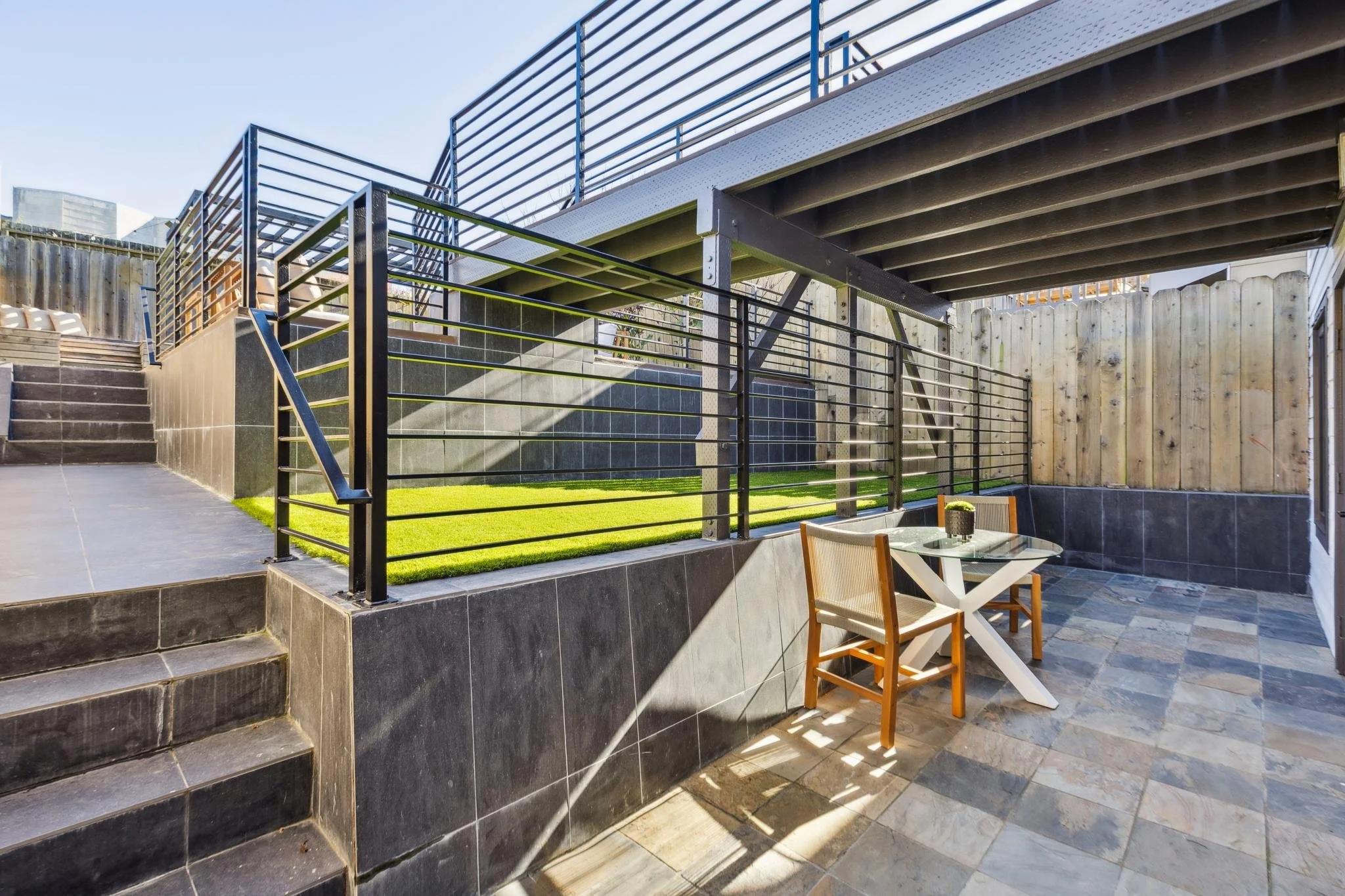 Outdoor patio area with black railings, a small grass patch, and tiled flooring. There's a glass table with two chairs and a small potted plant on the table. Wooden fence encloses the space, and construction beams support an upper deck.