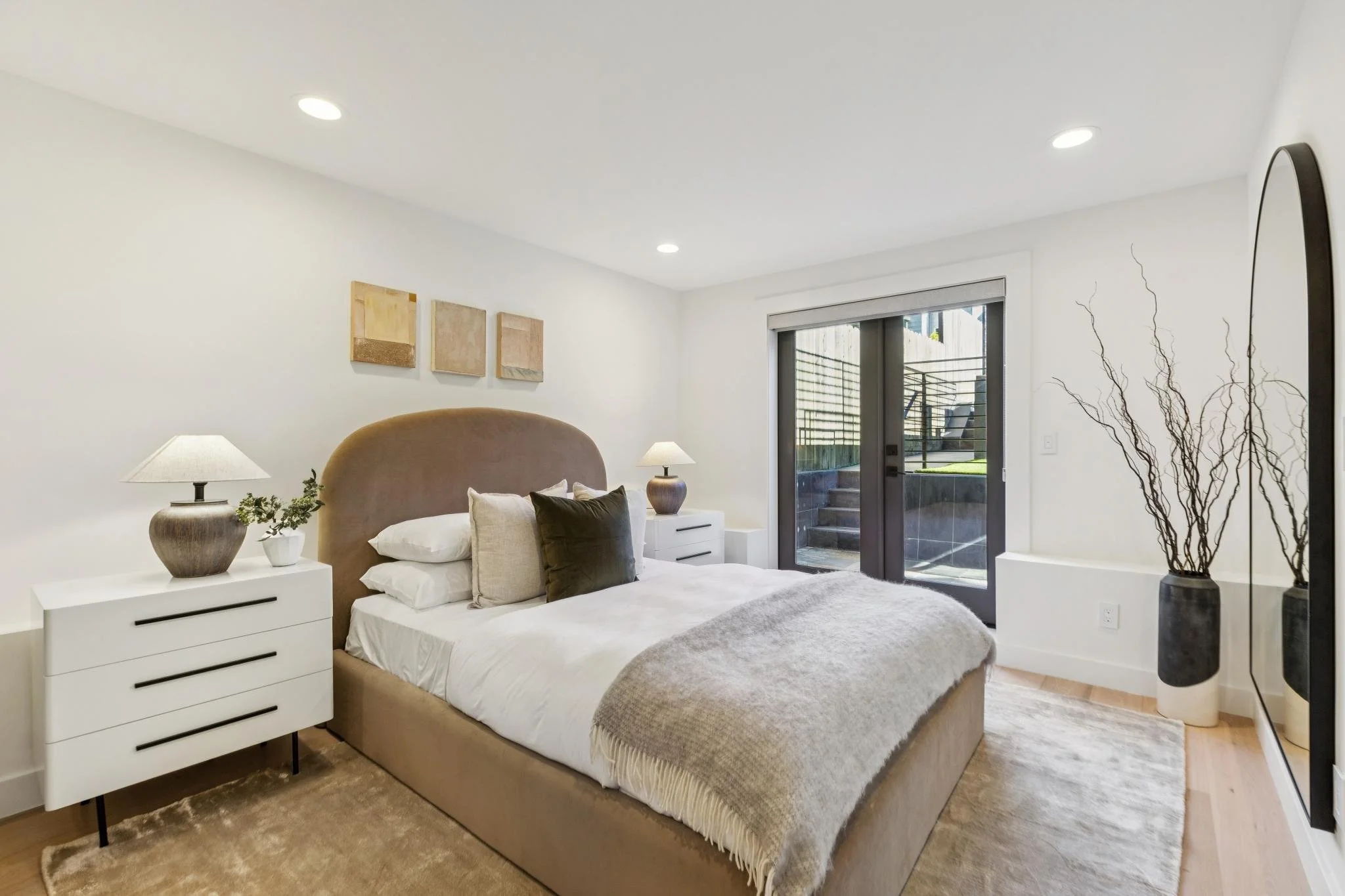 A modern bedroom with a beige upholstered bed, white bedding, and multiple pillows. Two white nightstands with lamps and decorative pieces are on each side of the bed. There are three abstract wall art pieces above the bed. A large mirror and tall va