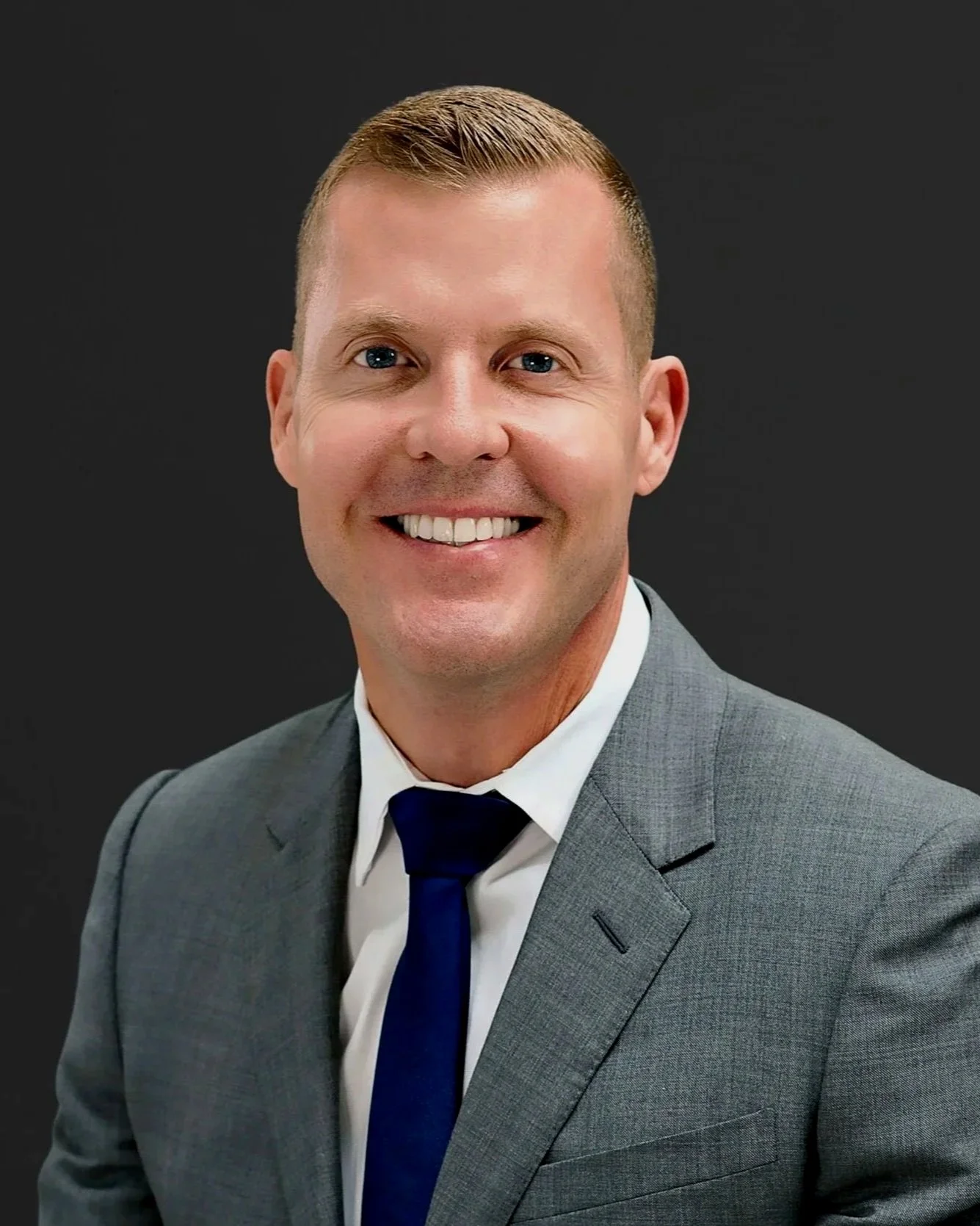 A man in a gray suit, white shirt, and navy tie smiling at the camera, against a dark background.